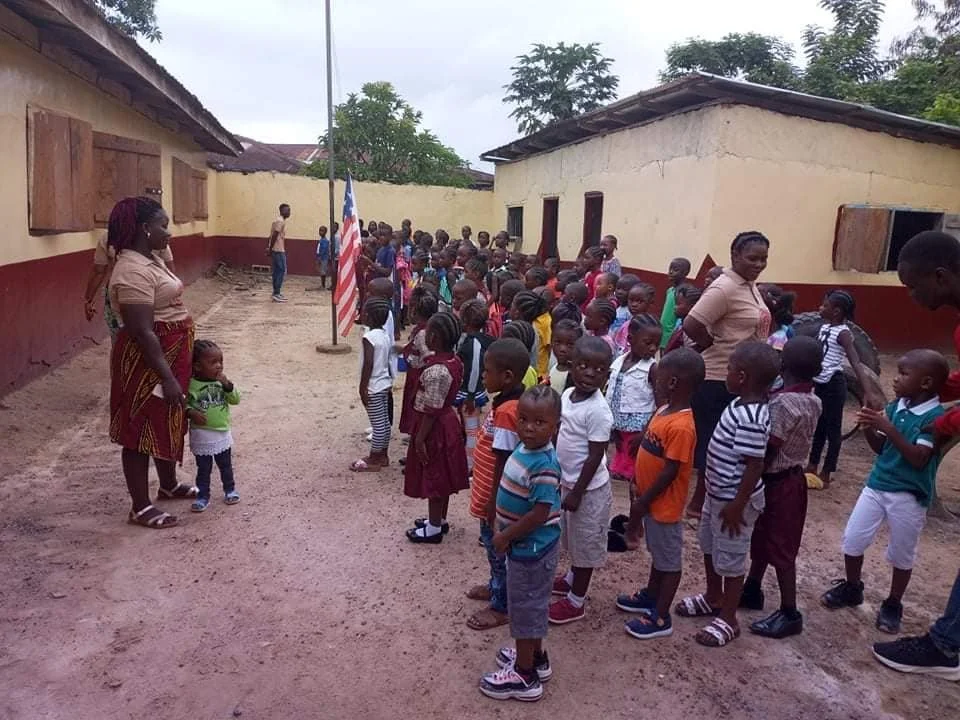 Children and teachers standing in lines outside of school buildings, with an American flag on a pole in an outdoor courtyard.