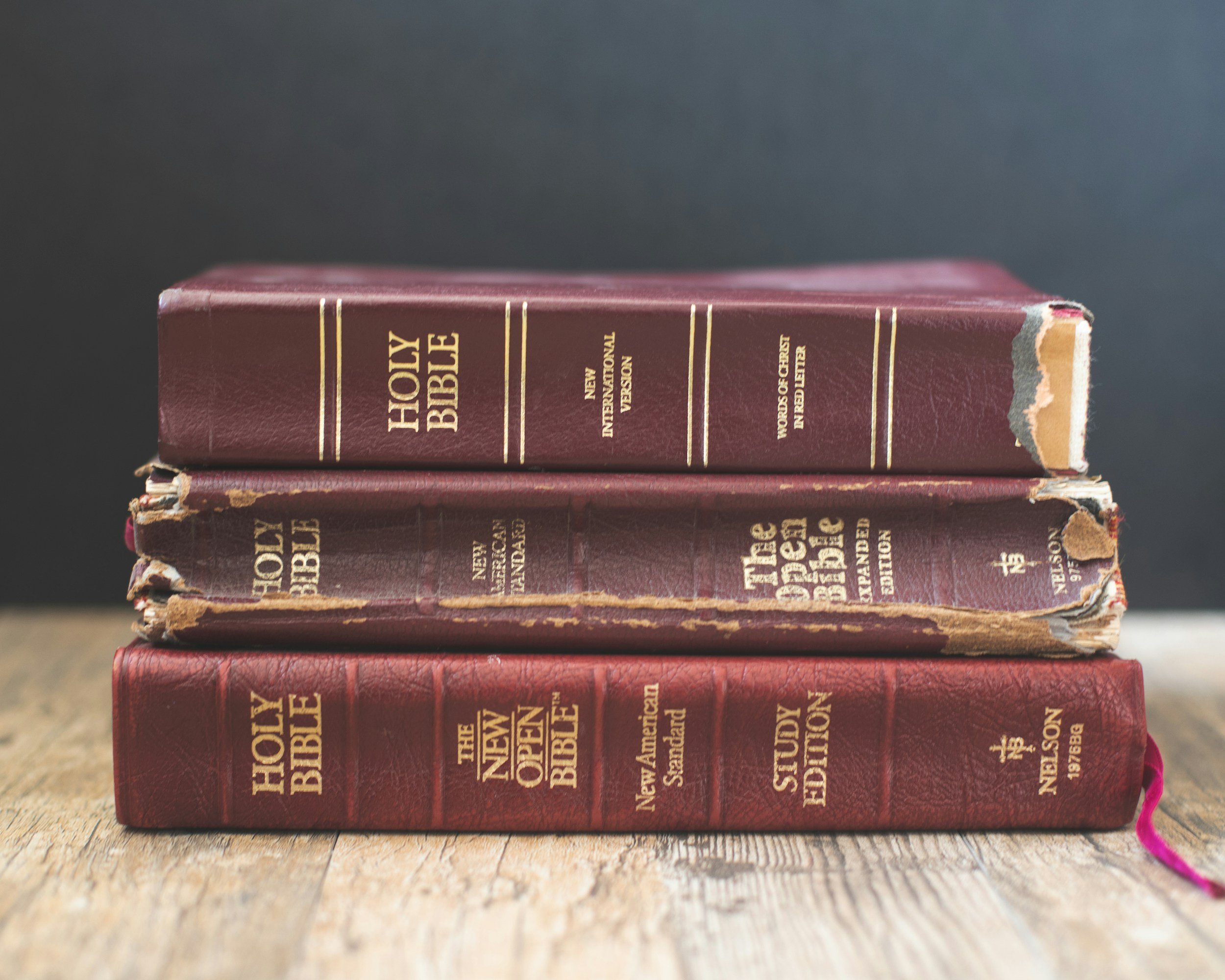 Three worn, hardcover Bibles stacked horizontally on a wooden surface, with the top Bible's spine visible, showing the title 'Holy Bible' in gold lettering. The middle and bottom Bibles also show the titles and have visible signs of wear and tear.