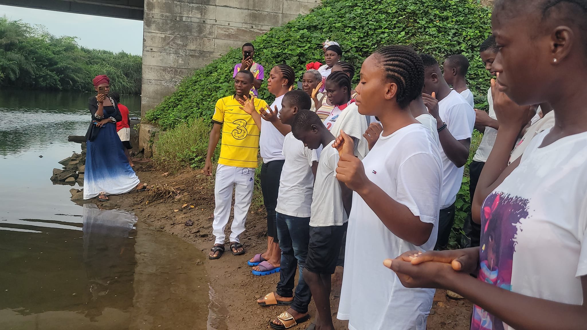 A group of people standing by the lakeside with a woman walking into the water under a bridge, some individuals with hands raised or clasped, engaged in a collective activity or prayer.