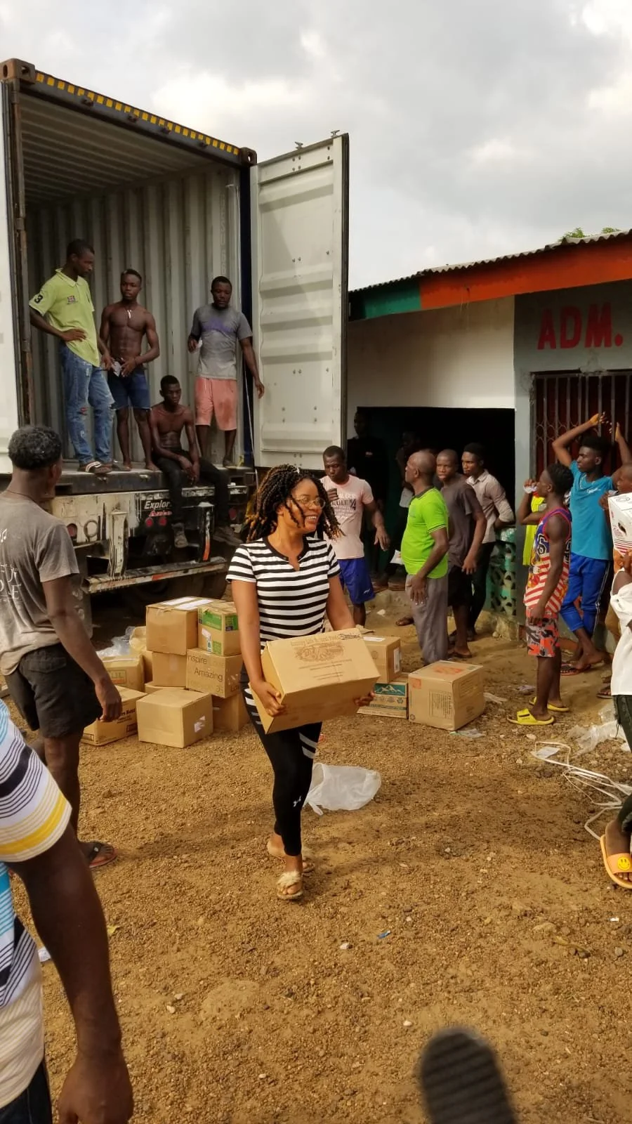 A group of people unloading boxes from a truck in an outdoor setting, with some standing inside the truck and others on the ground among the boxes. One woman in the foreground carries a box, smiling, wearing a black and white striped shirt and black pants.