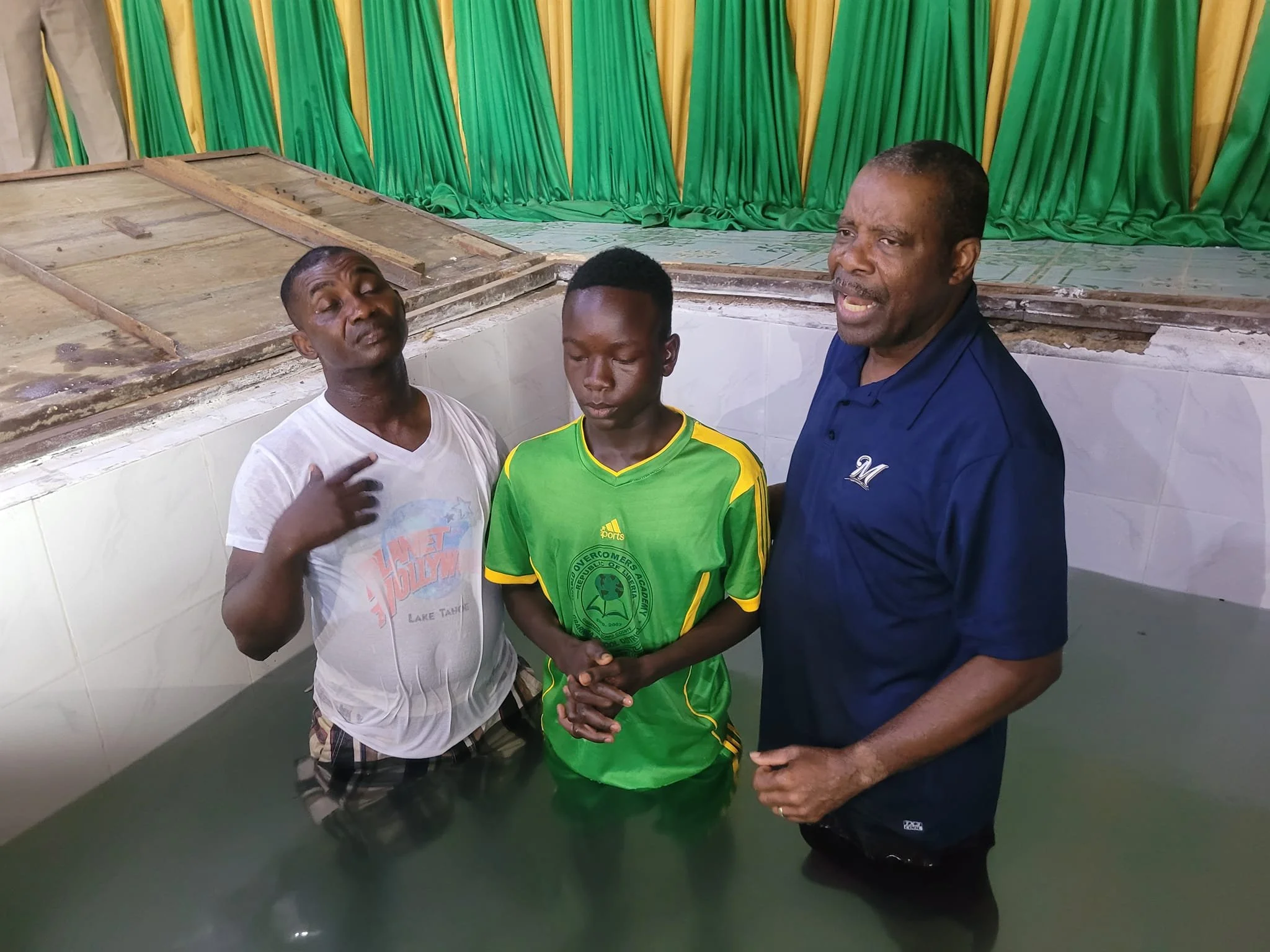 Three men standing in a tank of water indoors, with green and yellow curtains in the background. The man on the left is wearing a white t-shirt, the young man in the middle is wearing a green sports jersey, and the man on the right is wearing a dark blue shirt.