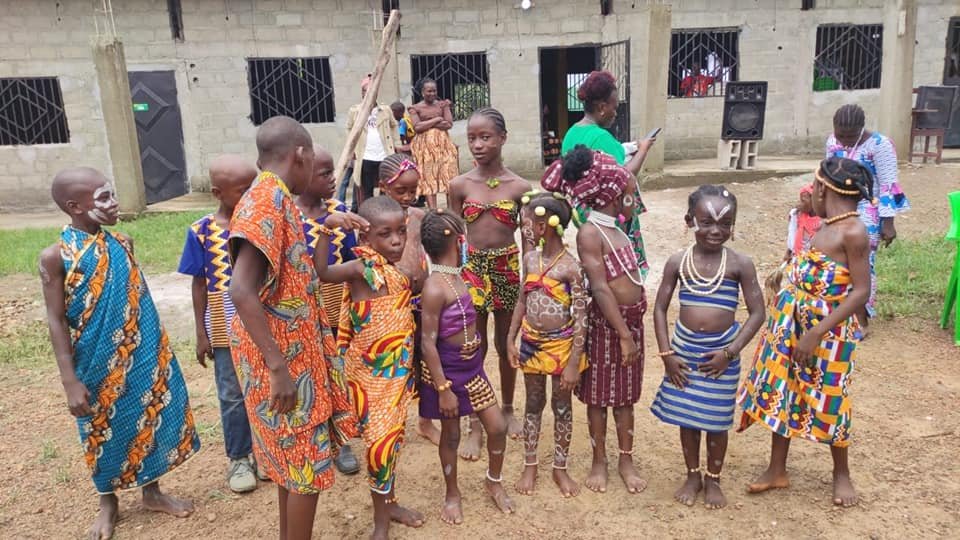 Group of children and women dressed in colorful traditional African clothing outdoors, with some children having face paint and jewelry, in front of a building with barred windows.