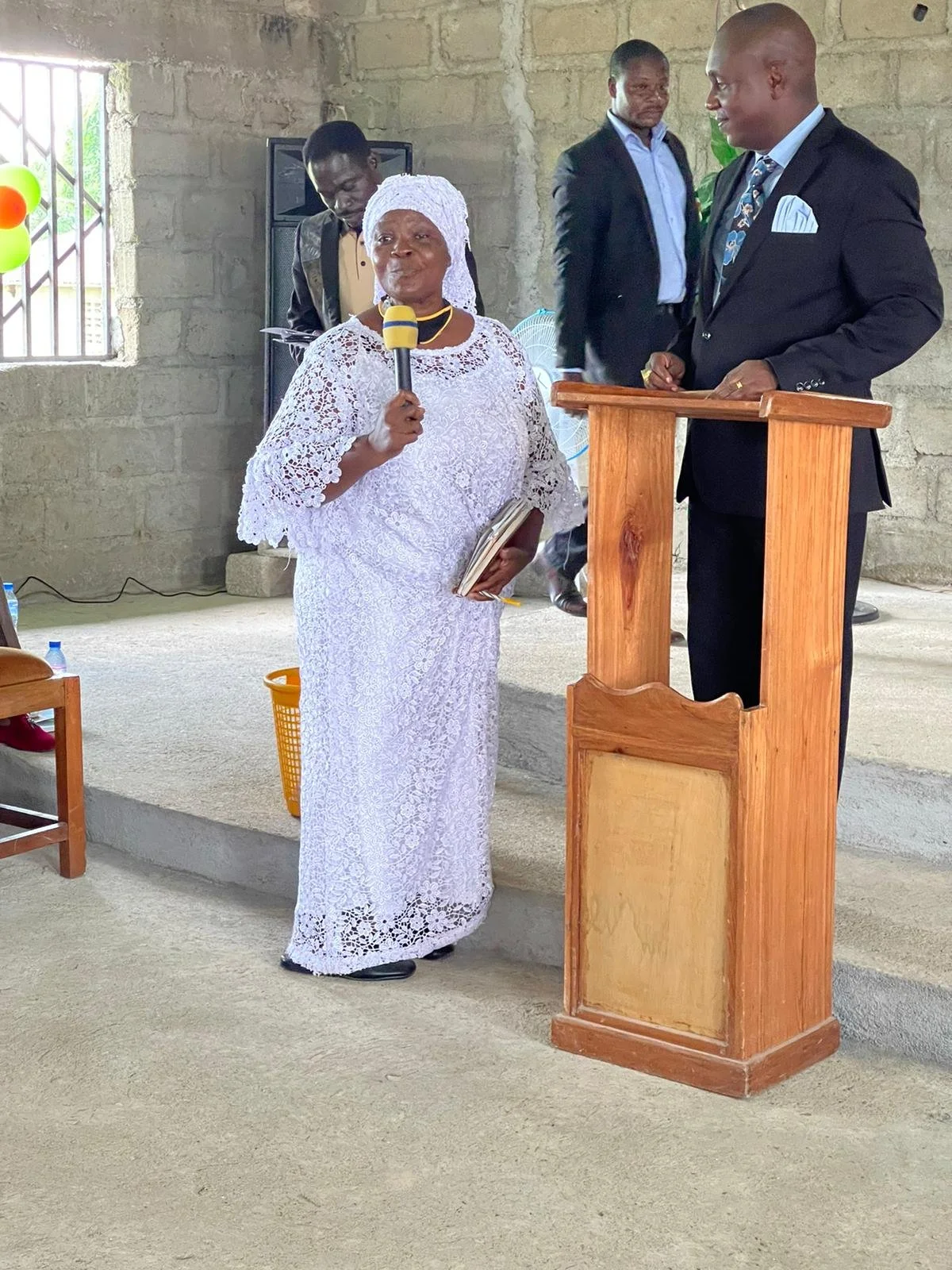 An elderly woman in white lace dress and headwrap speaking into a microphone in a church with men in suits listening nearby.