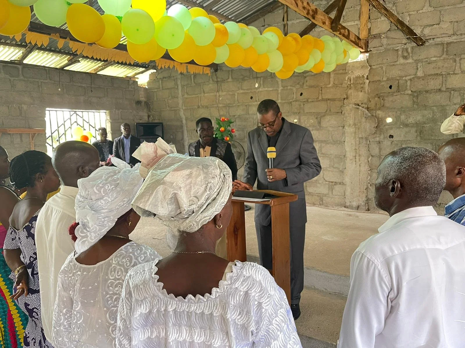 A group of people participating in a formal ceremony in an unfinished brick-walled room decorated with yellow balloons hanging from the ceiling. A man in a grey suit is reading from a book at a wooden podium, holding a microphone, while others stand and listen attentively, some dressed in traditional attire.