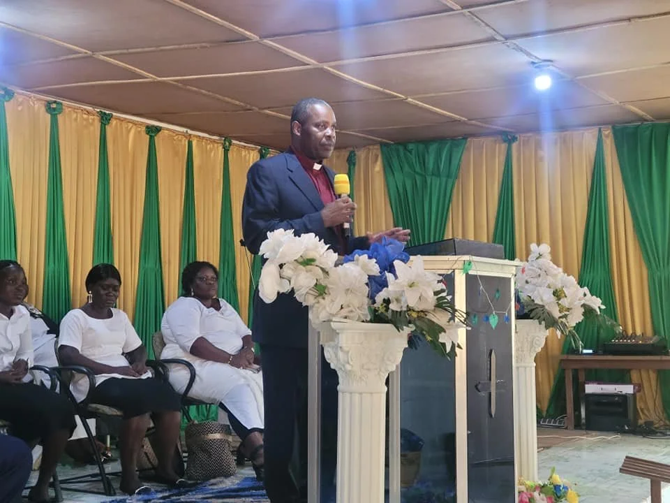 A man in a dark suit and maroon shirt stands at a podium decorated with white flowers, speaking into a yellow microphone. Three women in white attire are seated behind him, with green and yellow curtains as the backdrop in a room with a low ceiling.