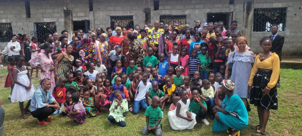 A large group of children and adults gathered outdoors in front of a concrete building, posing for a group photo on the grass field.