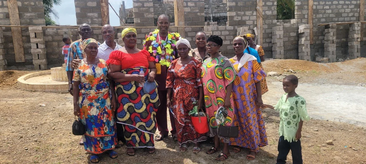 Group of people posing in front of a building under construction, including men and women wearing colorful traditional clothing and a young boy in a light green shirt.