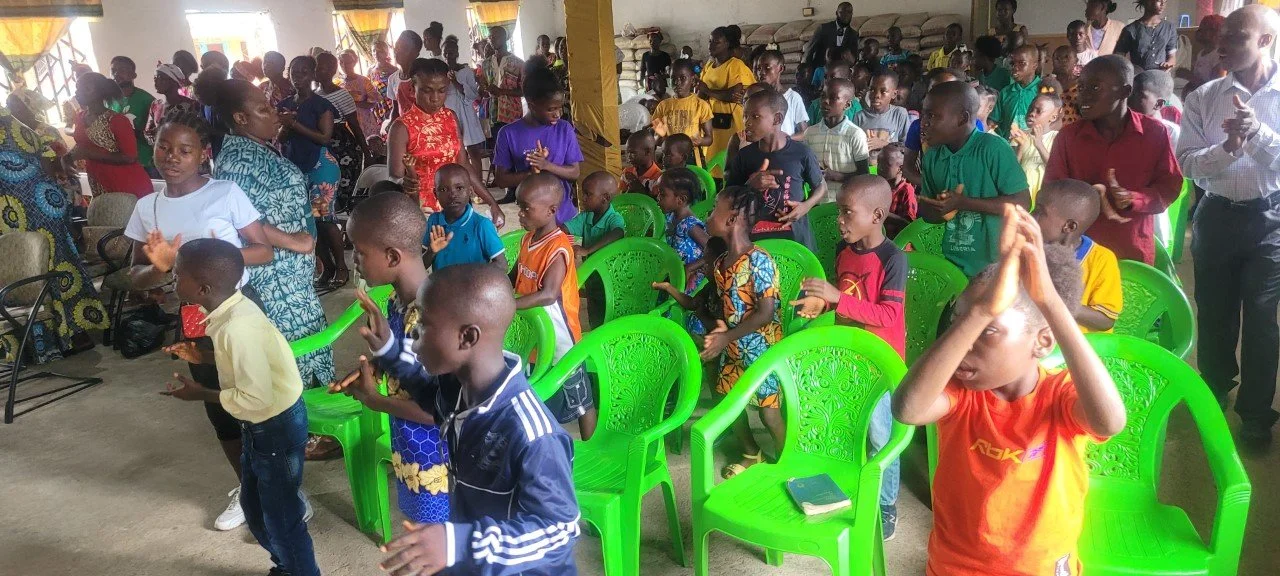 Children and adults gathered in a hall, some seated and some standing, participating in a group activity or event. Children in the front are actively engaged, some with hands raised or clapping.