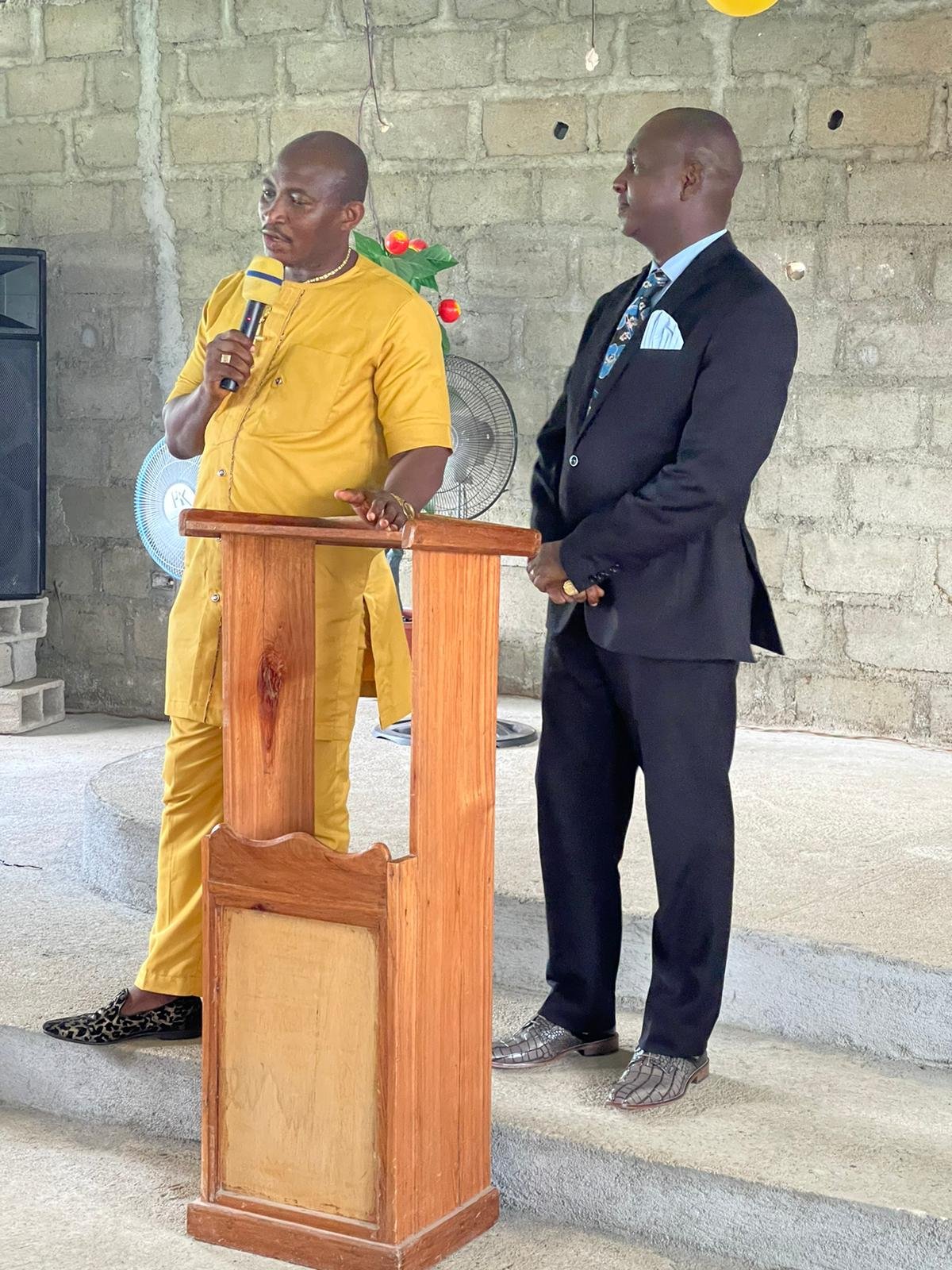 A man in a yellow outfit is speaking into a microphone at a wooden podium, while another man in a black suit with a blue shirt and patterned tie stands beside him in an indoor setting with a brick wall background.