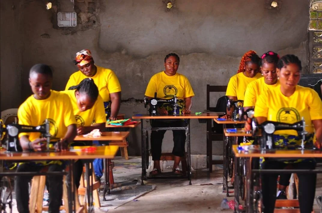 Women, some children, in yellow shirts working on sewing machines in a rustic room.