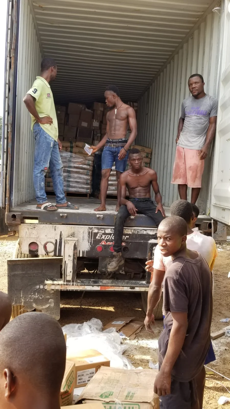 A group of young men standing and sitting inside the back of a large shipping truck, with stacked boxes and packages around them, in an outdoor setting.