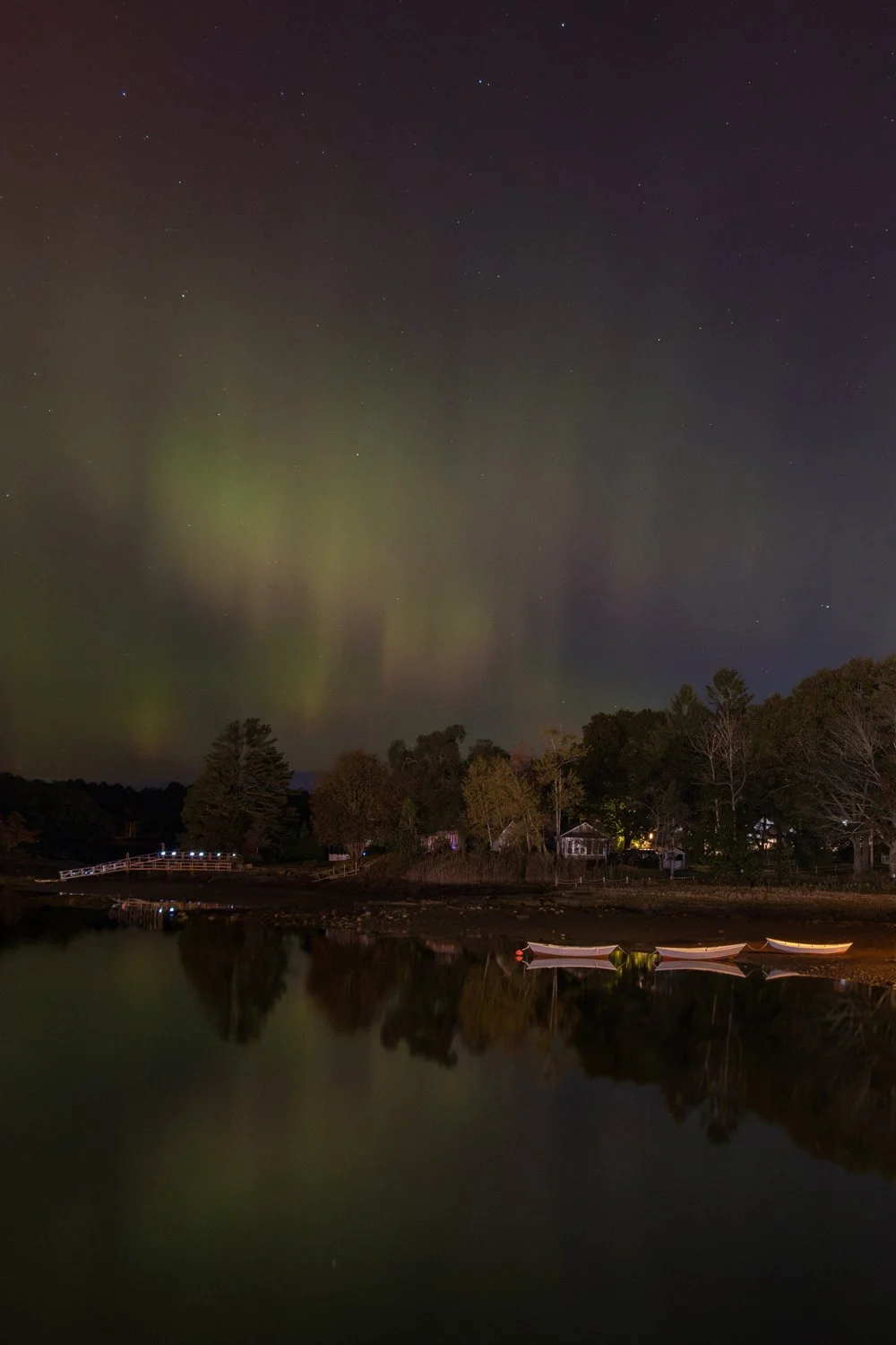 Nighttime scene of the northern lights over the coast of Kennebunkport Maine with small boats and a wooded shoreline.