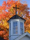 Small church steeple with a cross on top against autumn trees with orange leaves