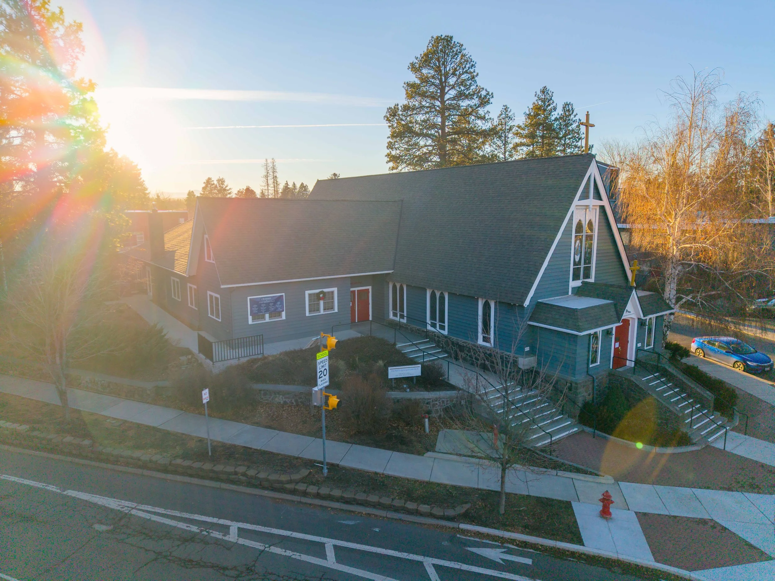 A church building with blue siding and white trim, steep roof, cross on top, located on a street corner during sunset with trees around.