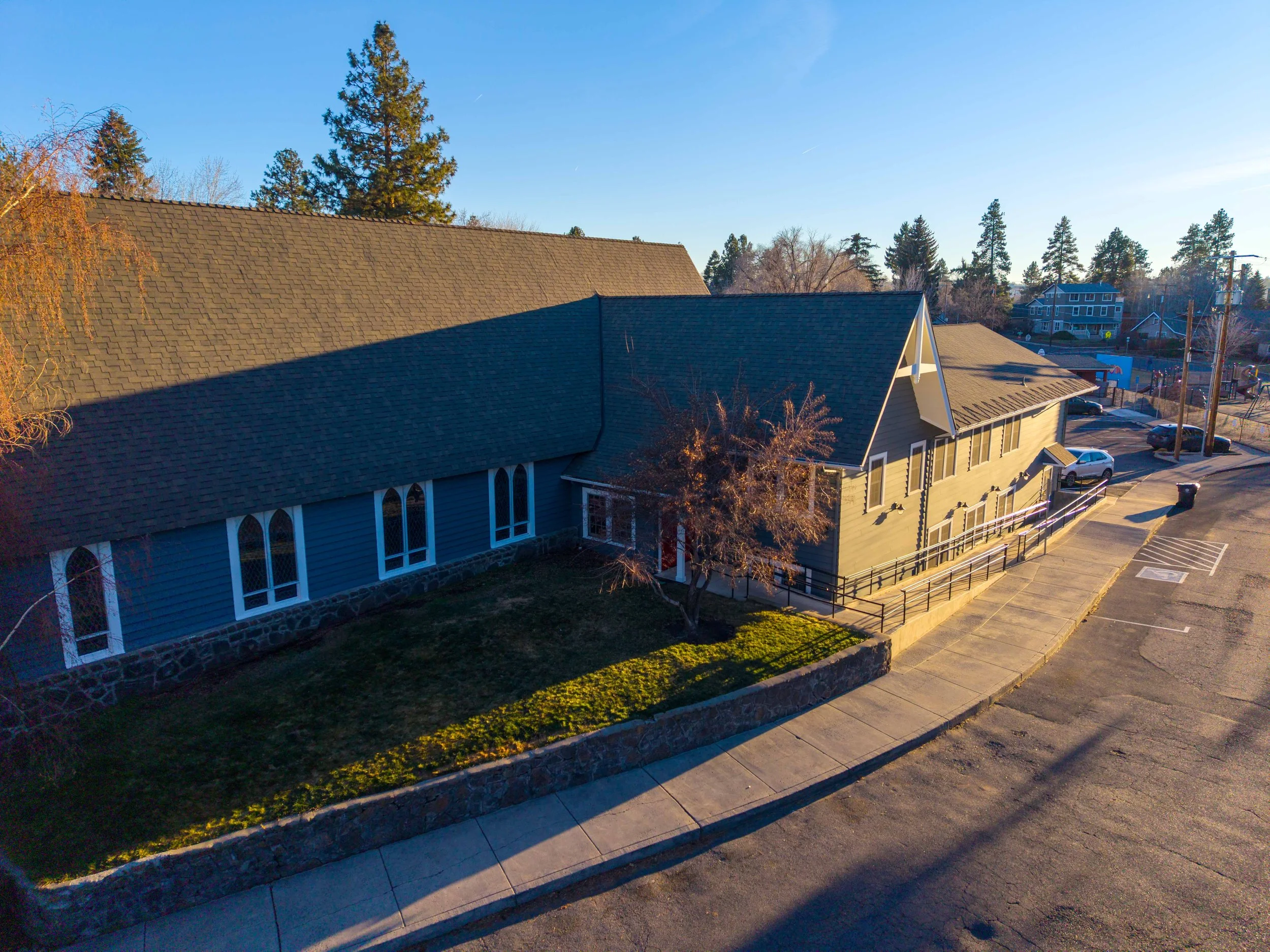 A church building with a blue exterior, large arched stained glass windows, and a steep roof, surrounded by a small lawn and trees, with a walkway and parking lot in front, during clear weather.