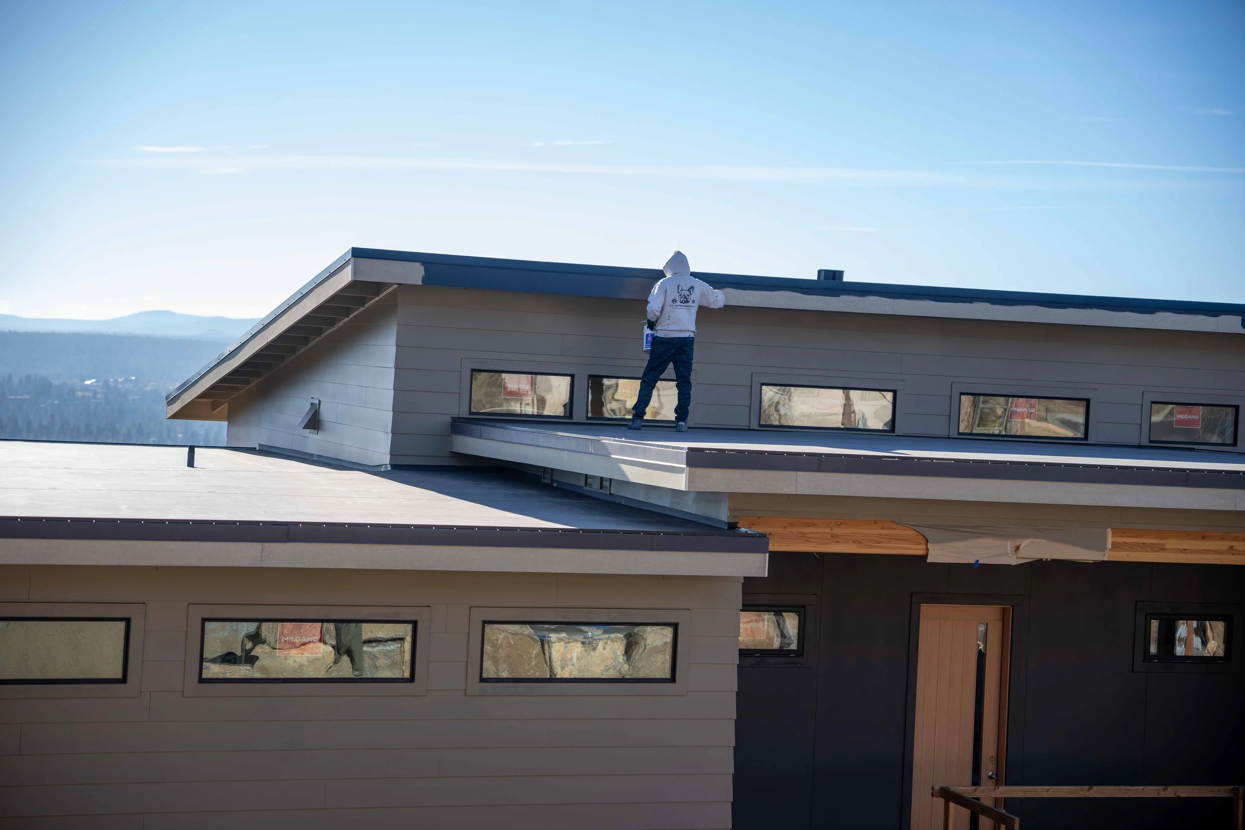 A construction worker in a white hoodie stands on the roof of a modern house with angled rooflines and multiple windows, with scenic mountains in the background.