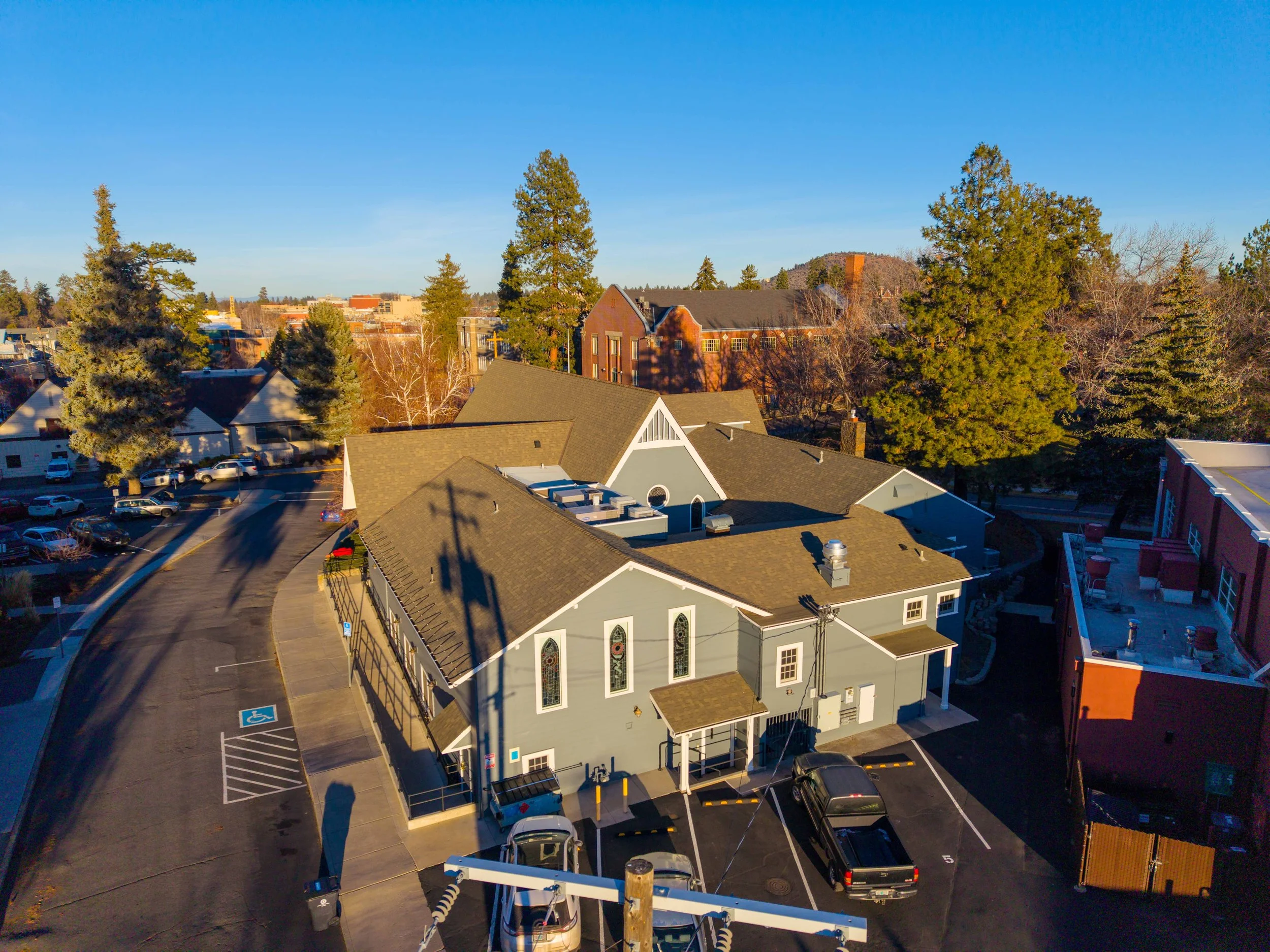 Aerial view of a church building with a parking lot, situated in a town with trees and additional buildings in the background, under a clear blue sky.