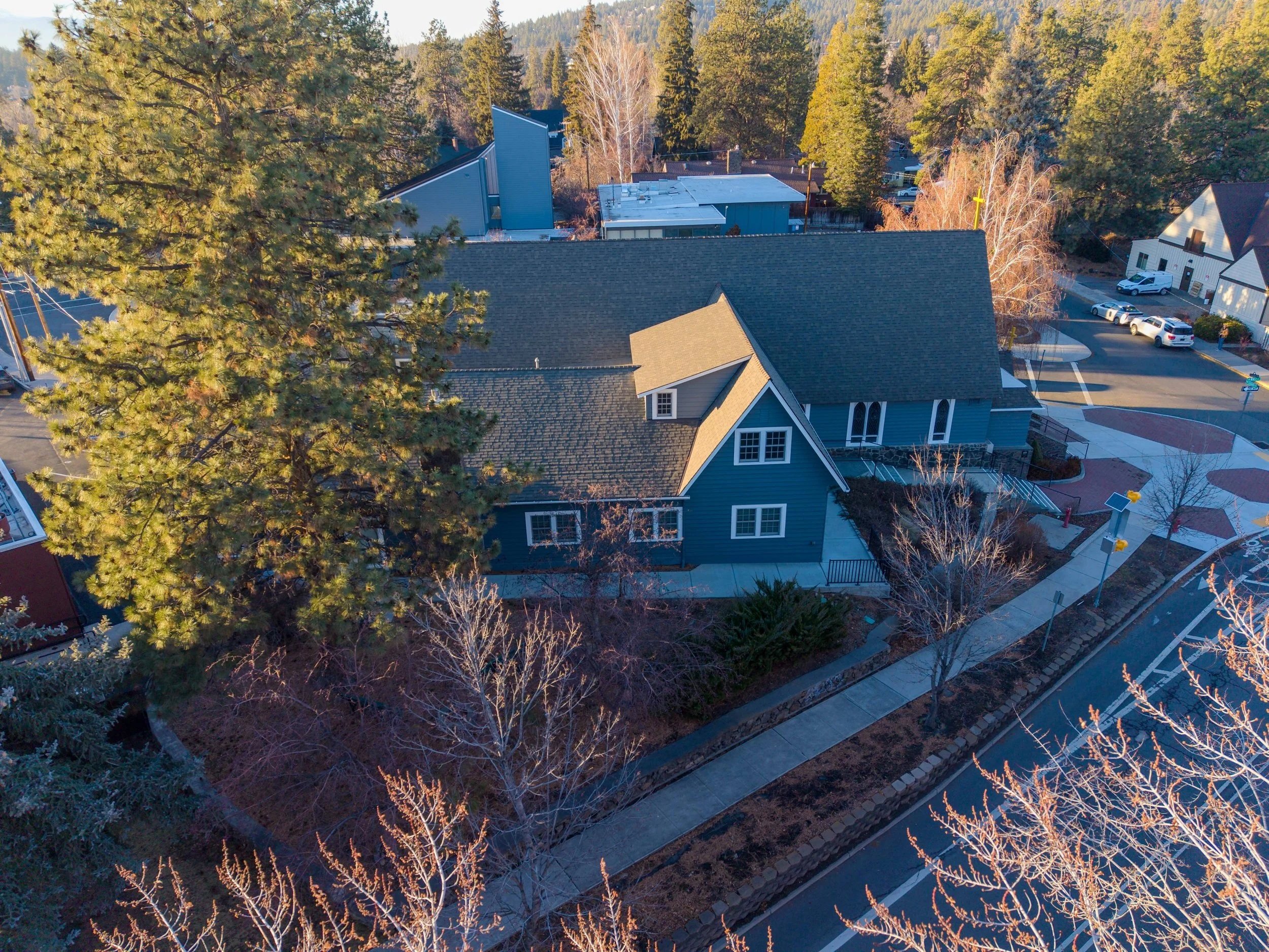 An aerial view of a house in a neighborhood, surrounded by trees and parked cars, with a sidewalk and street in the foreground.