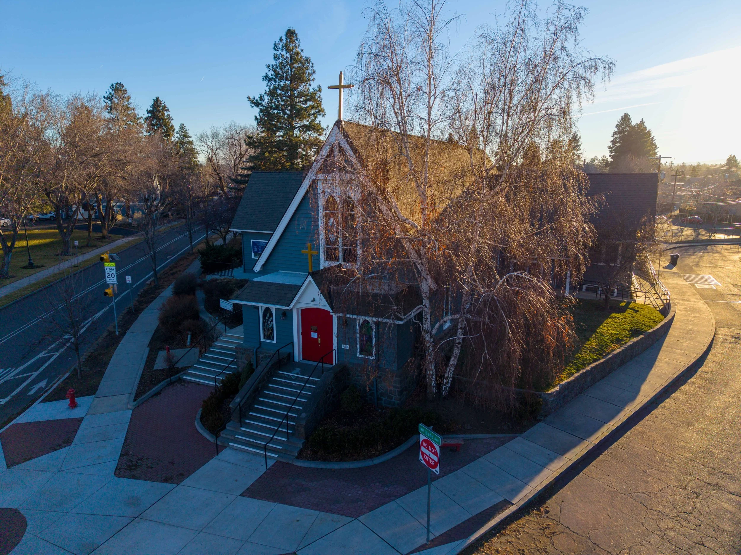 An aerial view of a small church with a blue exterior, white trim, and a red door, surrounded by a sidewalk and trees, with a cross on the roof and a winding sidewalk leading to the entrance.