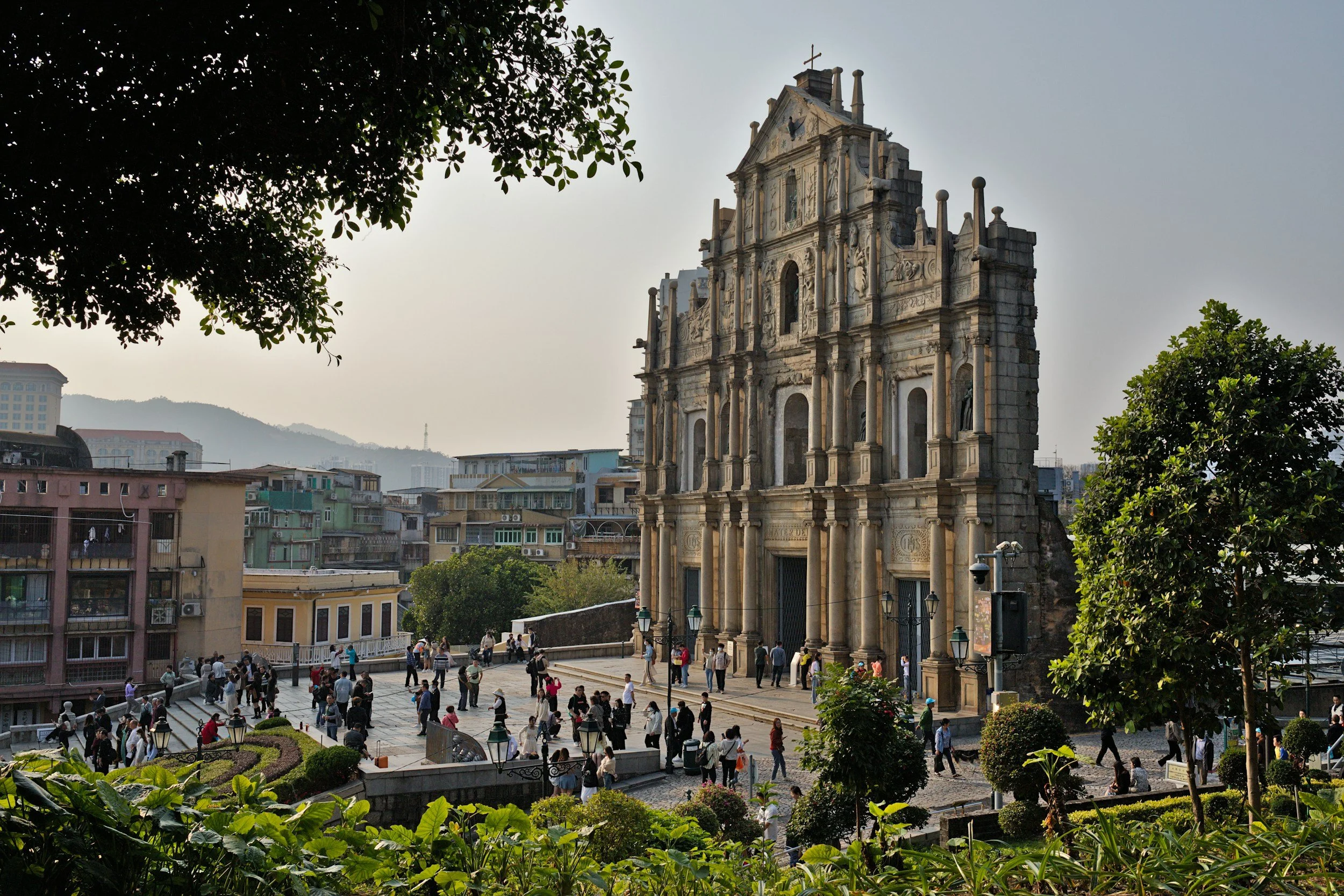 Historic church made of stone with elaborate architectural details, situated on a lively square filled with people, surrounded by trees and urban buildings.