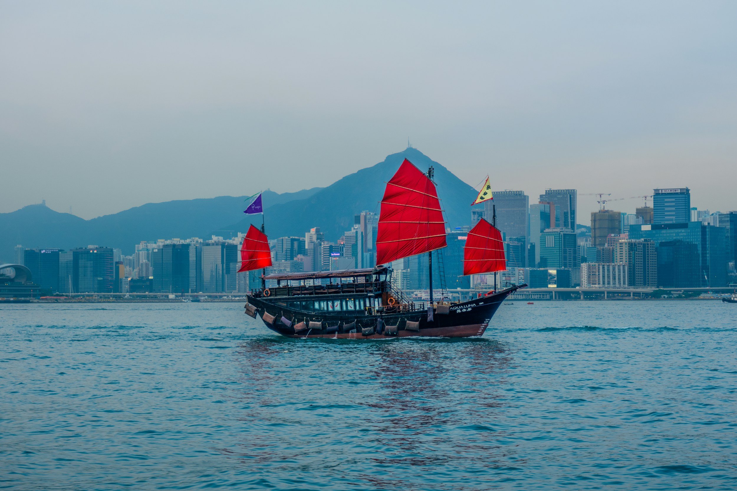 A traditional pirate-style ship with red sails sailing on the water near a modern city skyline with tall buildings and mountains in the background.