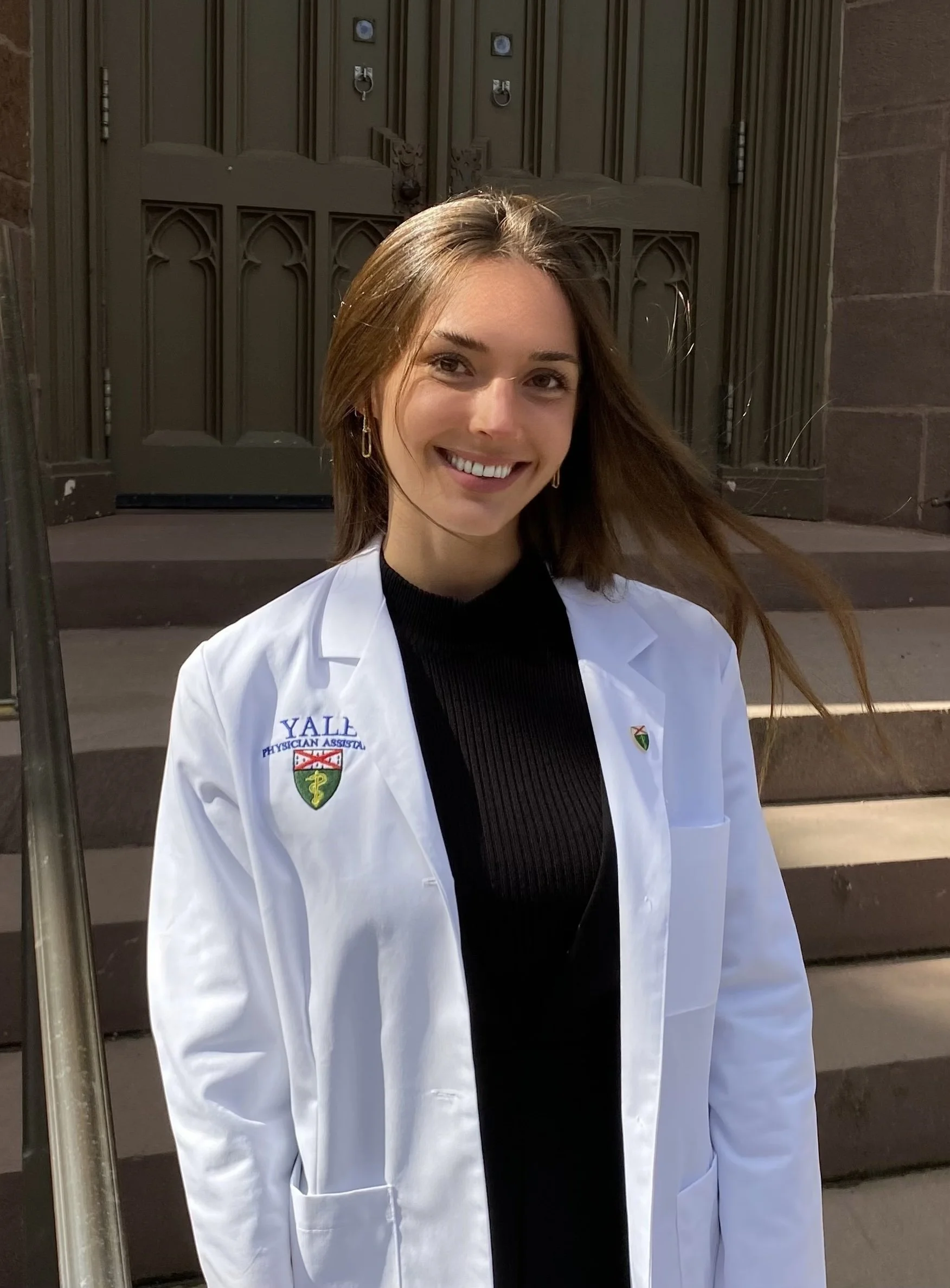 Young woman with brown hair smiling, wearing a white Yale physician assistant coat, standing on steps outside a building with a large wooden door.