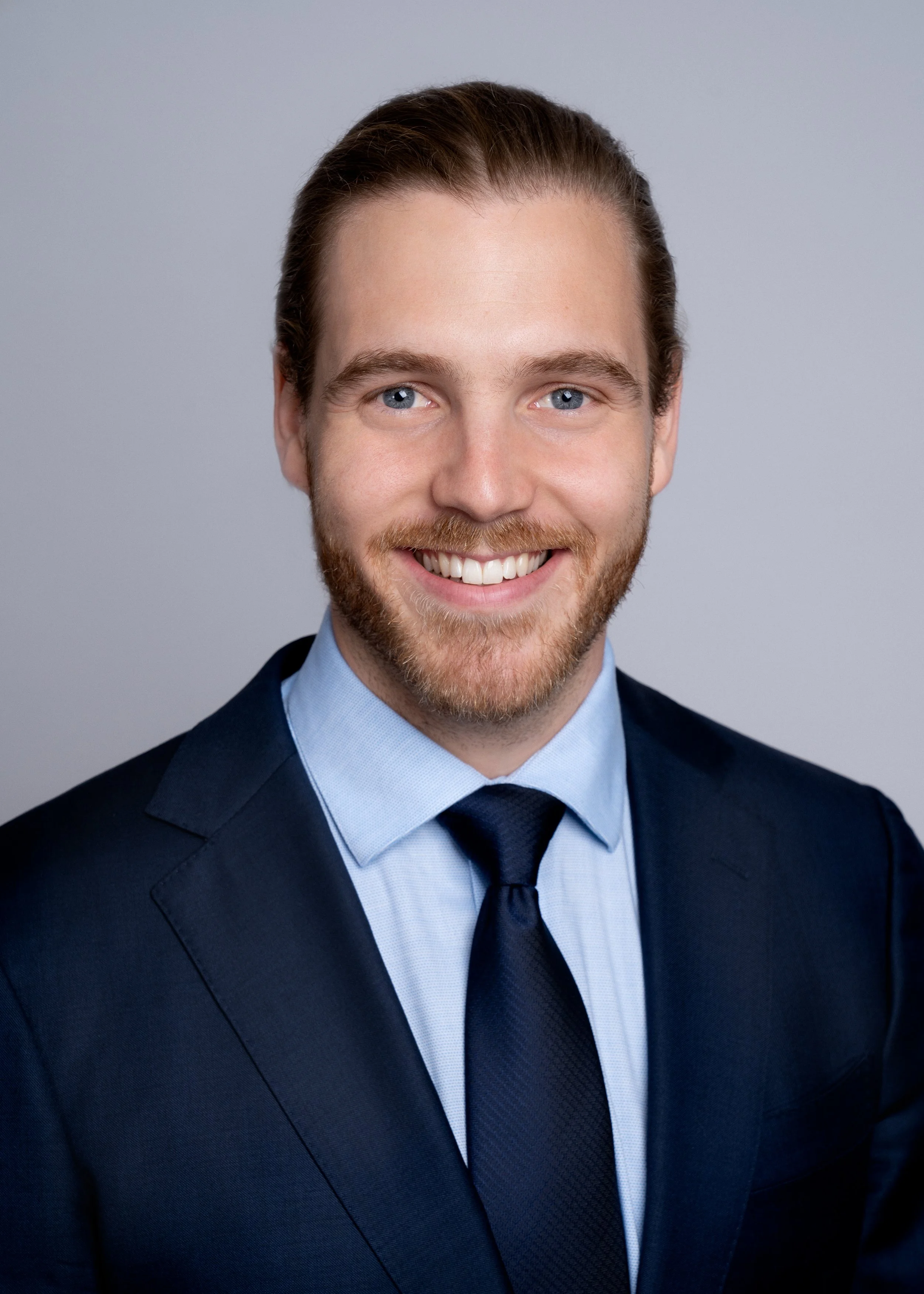 A headshot of a young man with light brown hair, a beard, wearing a dark suit, light blue shirt, and a dark tie, smiling against a plain gray background.