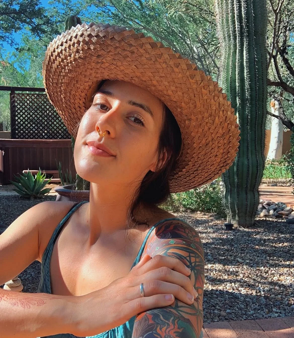 A woman wearing a large straw hat, sitting outdoors with desert plants and cacti in the background, smiling and looking at the camera.