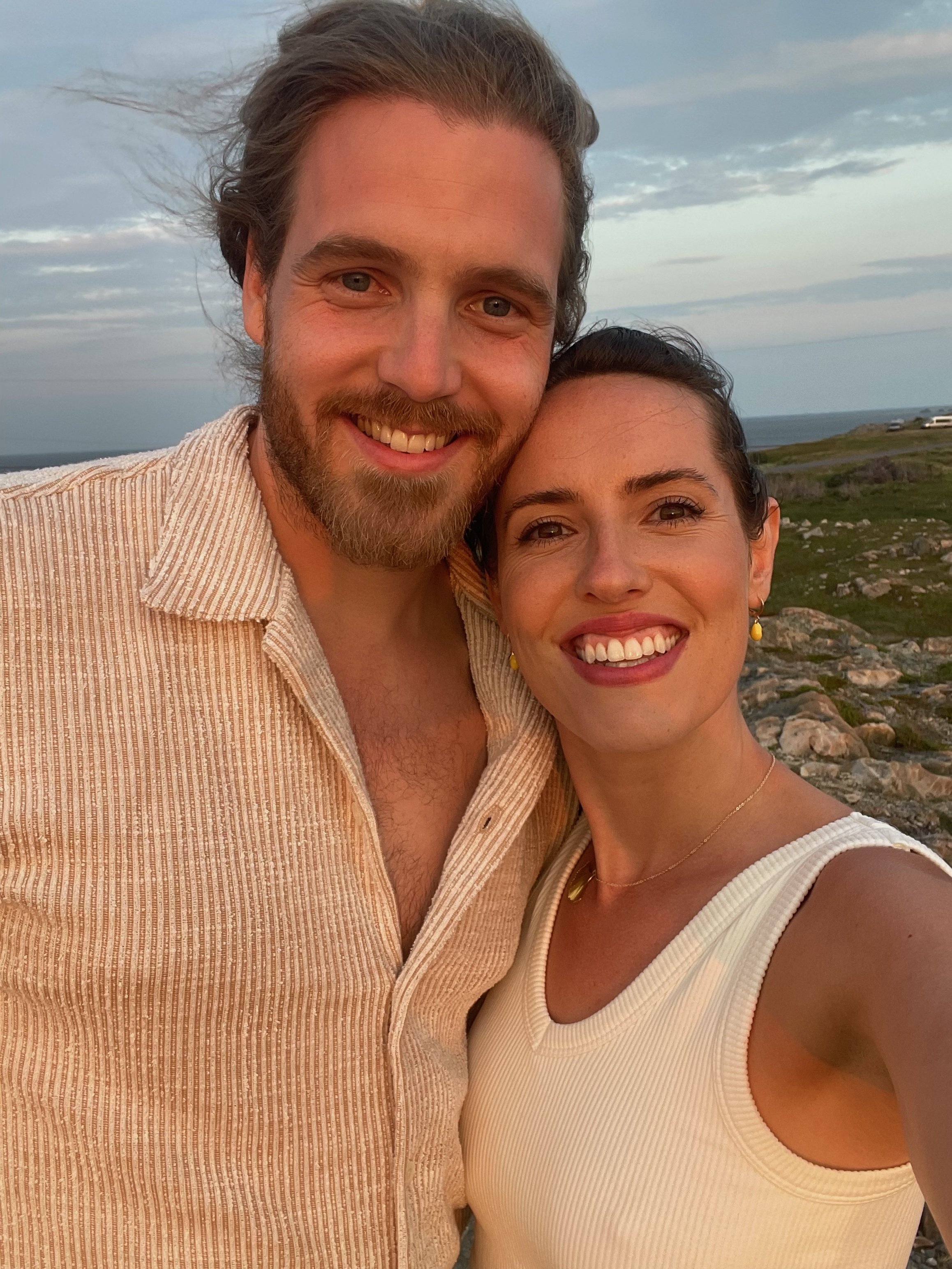 A smiling man and woman taking a selfie outdoors with a rocky landscape and cloudy sky in the background.