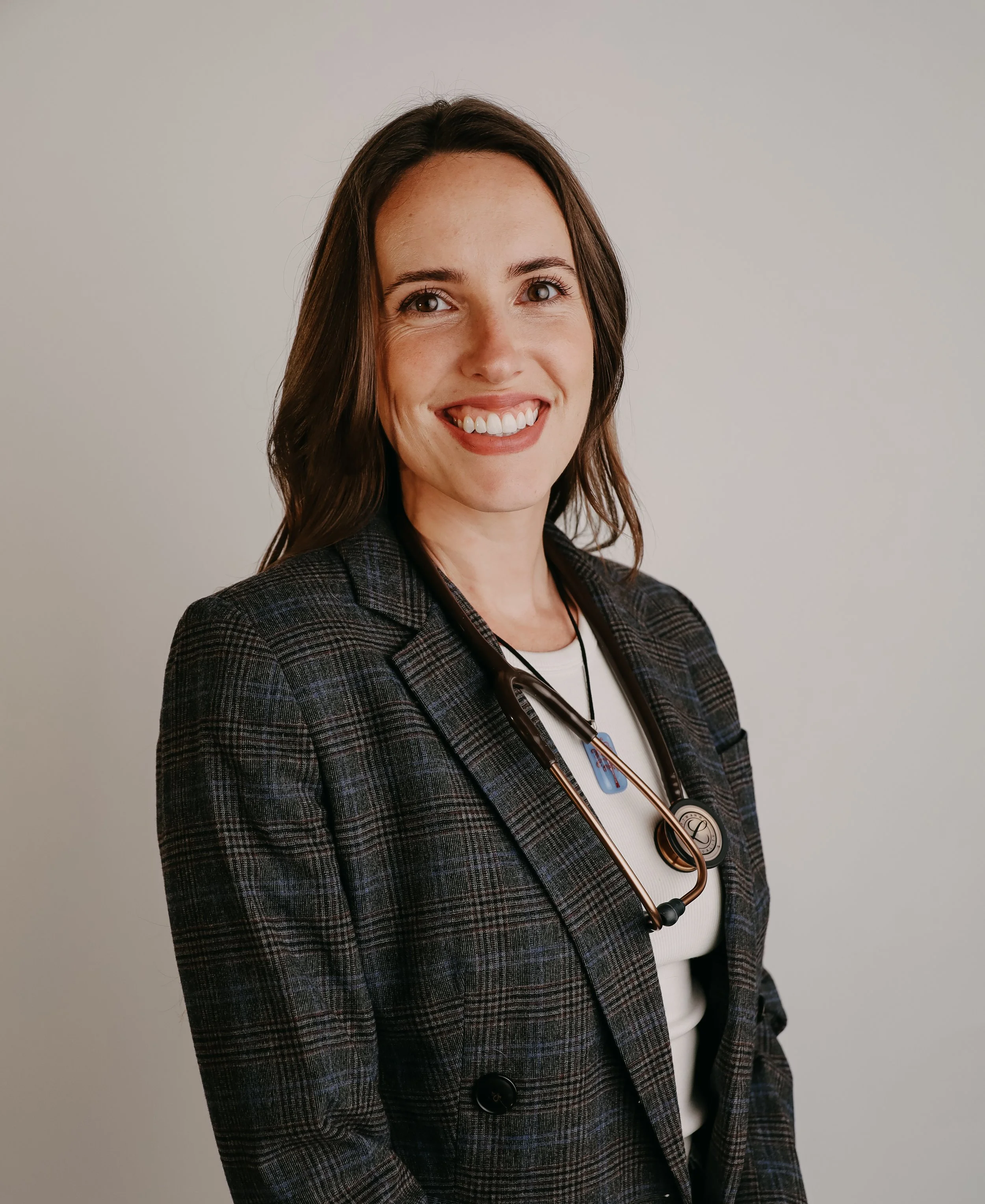 A smiling woman with brown hair wearing a plaid blazer and a stethoscope around her neck.