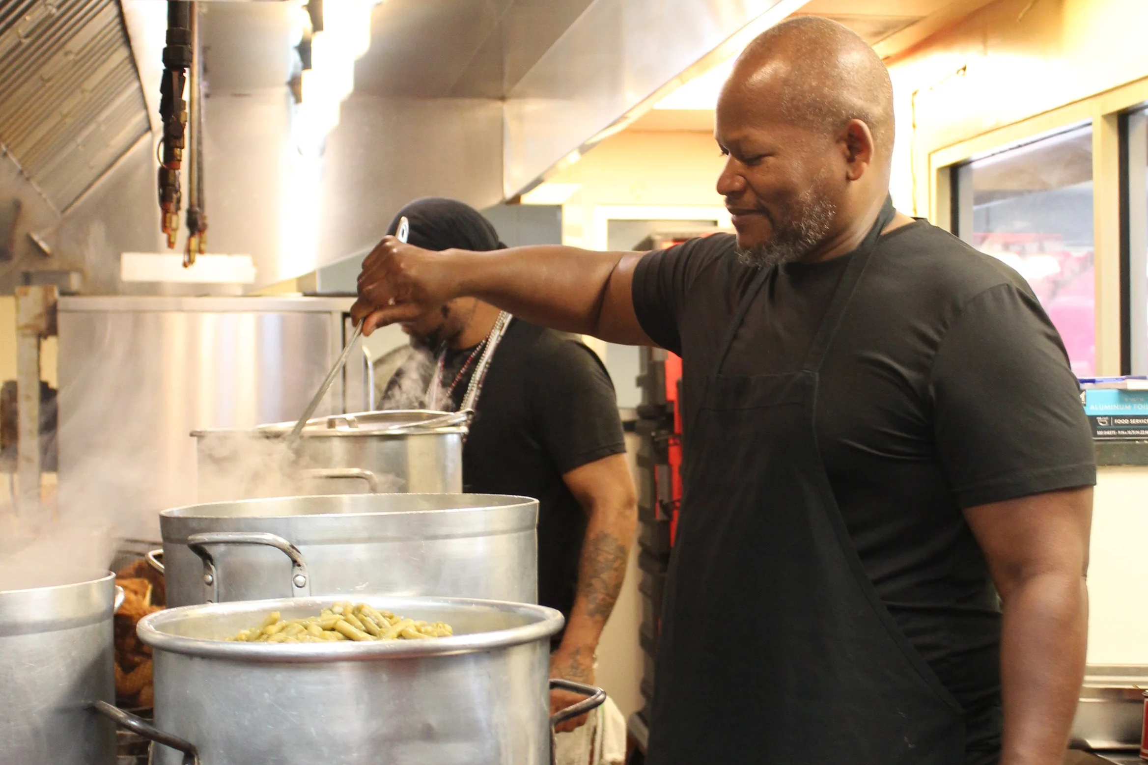 A man with a shaved head and gray beard stirring food in a large pot at a restaurant kitchen.