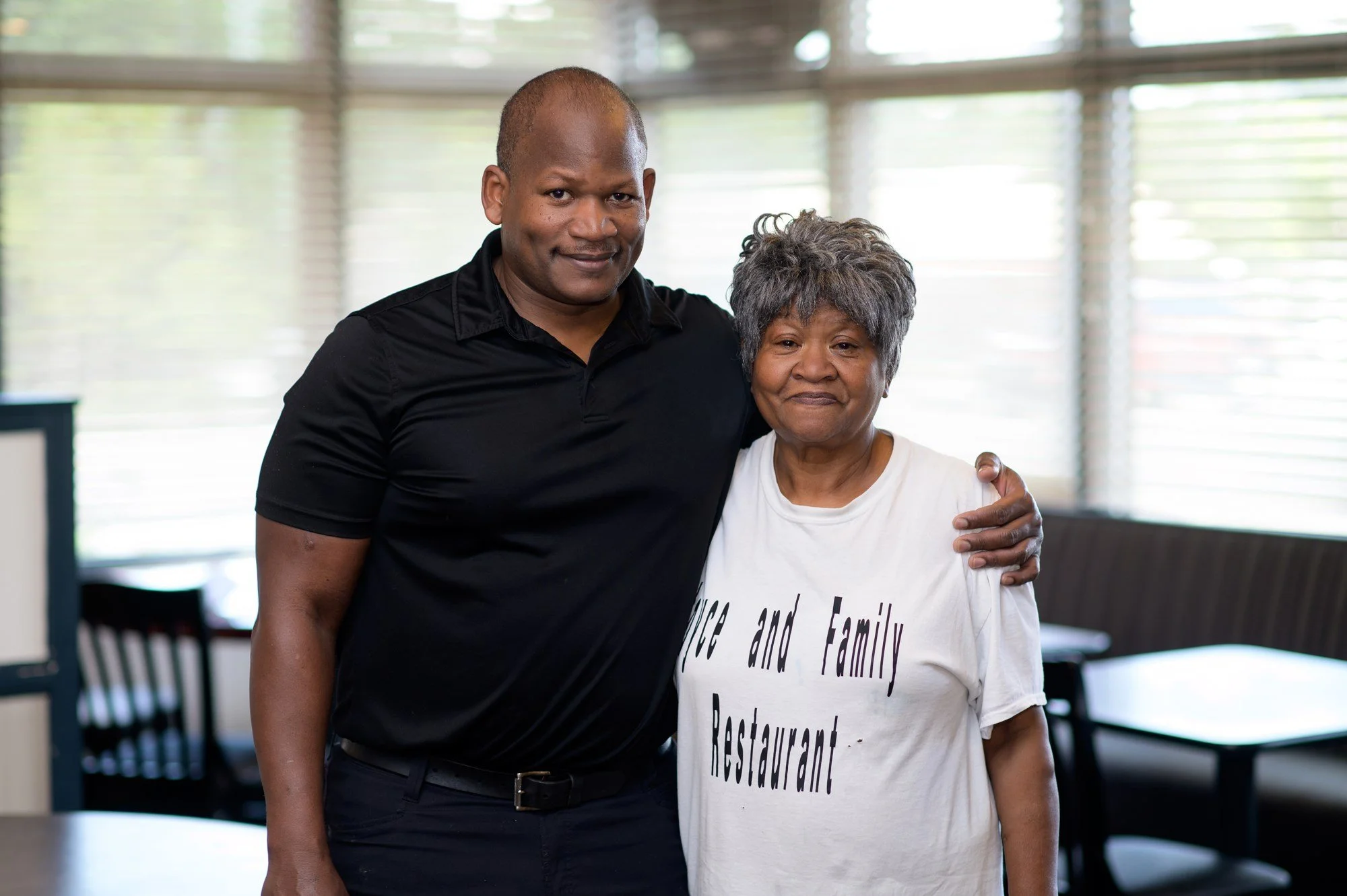 A man and an older woman standing together indoors, with the man having his arm around the woman. The woman is wearing a white T-shirt with black text, and the man is dressed in a black shirt. They are smiling gently at the camera in a room with large windows and blinds in the background.