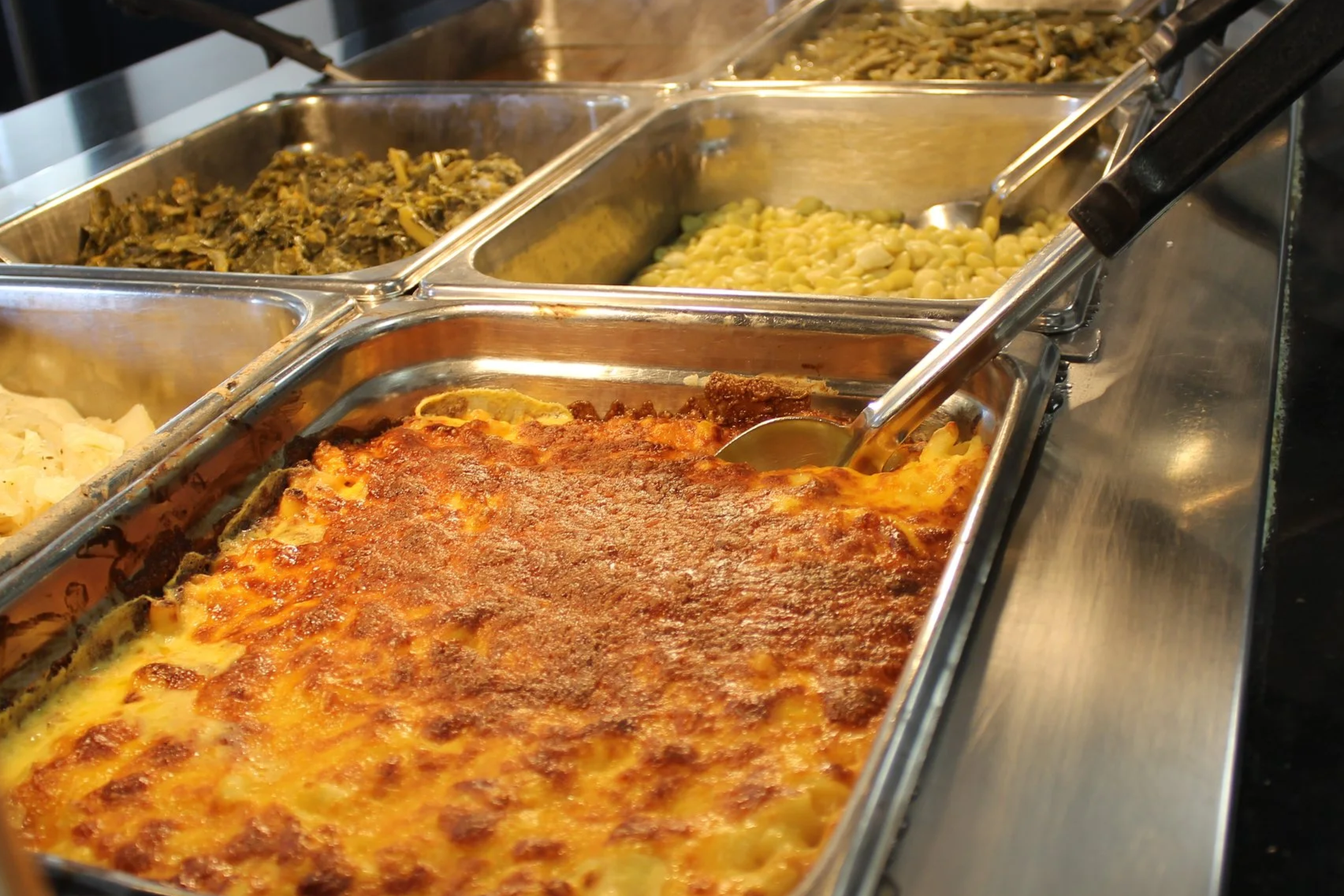 Buffet trays filled with baked pasta, cooked greens, macaroni and cheese, and mashed potatoes.