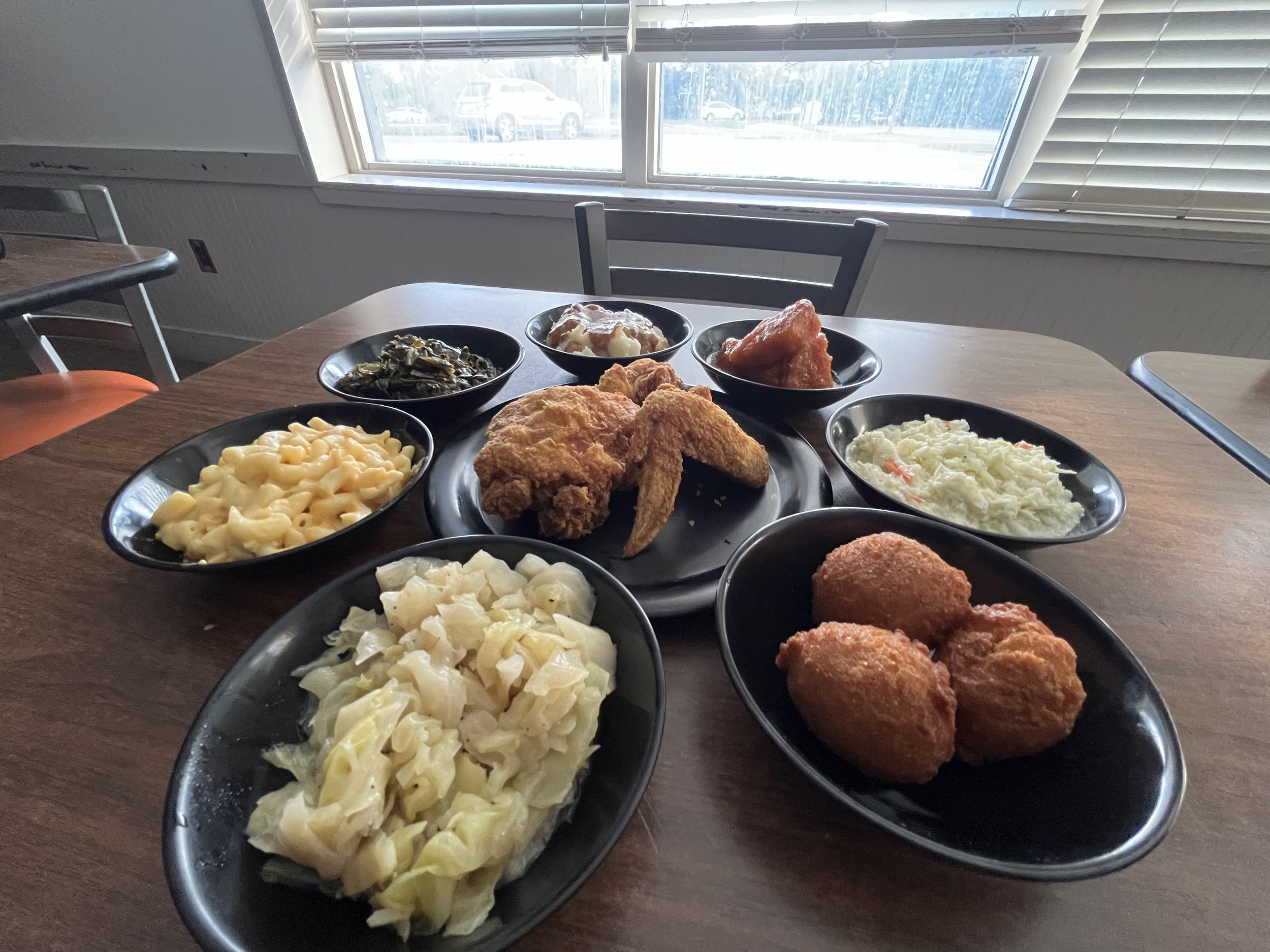 A table with several black plates filled with food, including macaroni and cheese, coleslaw, fried chicken, and other vegetables. In the background, a window with blinds shows a parking lot with several cars outside.