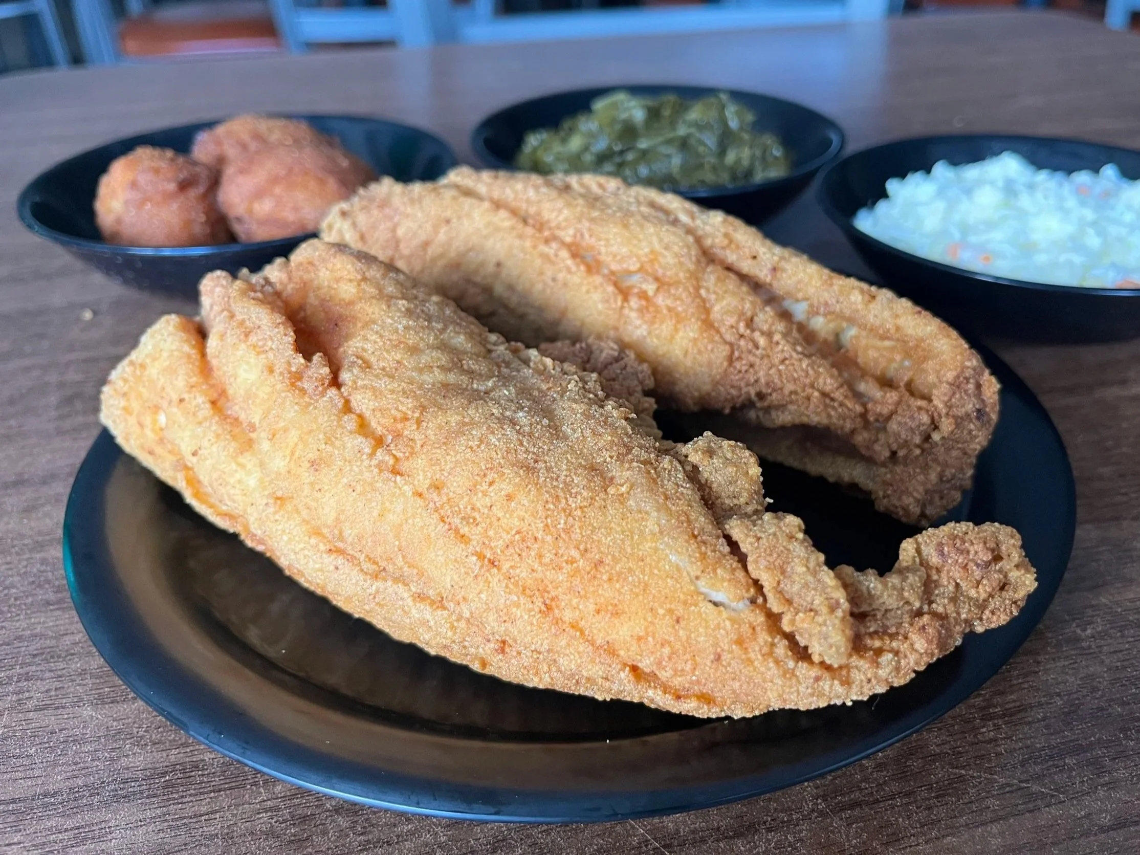 Fried fish fillets served on a black plate with sides of coleslaw, sautéed greens, and fried potatoes.