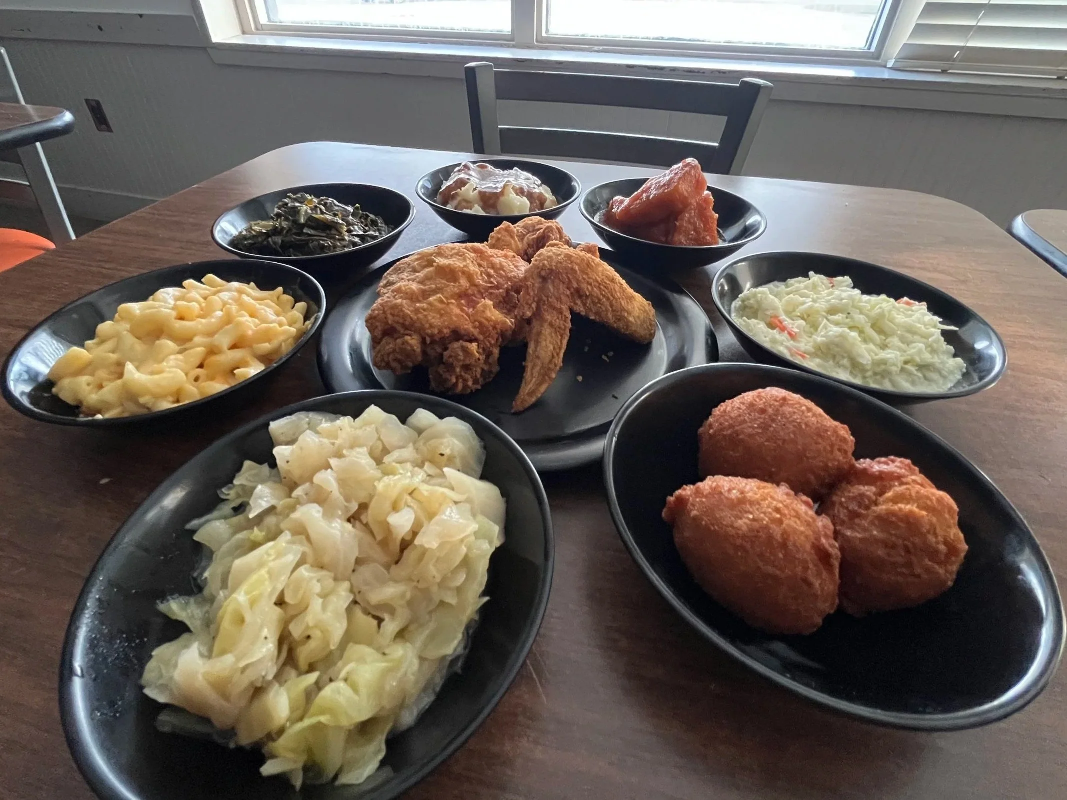 A table with various plates of food, including macaroni and cheese, fried chicken, mashed potatoes with gravy, cooked greens, sauced chicken, potato salad, and hush puppies. There is a window in the background and a chair is visible.