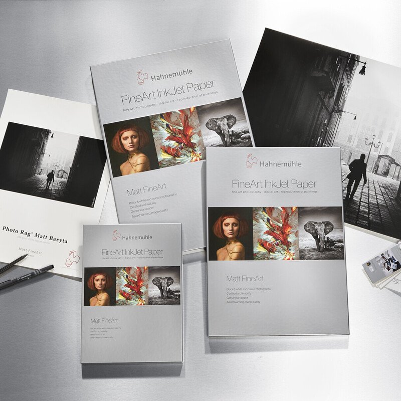Boxes of Hahnemühle FineArt InkJet Paper alongside black and white artwork prints of a woman, an elephant, and a street scene with silhouettes.}
