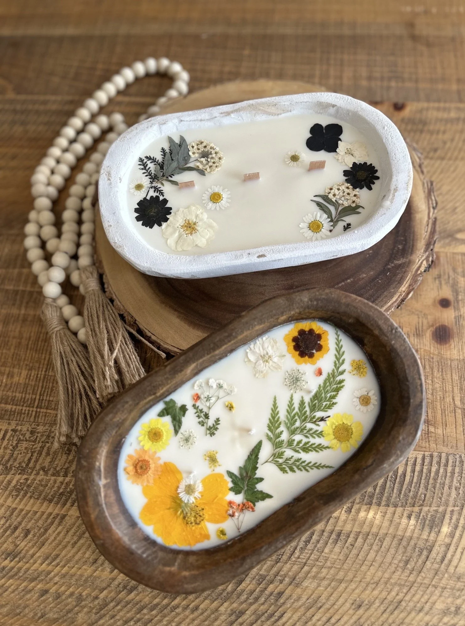 Two decorative candles with floral arrangements floating on white wax, placed on wooden coasters on a wooden surface, with a beaded necklace nearby.