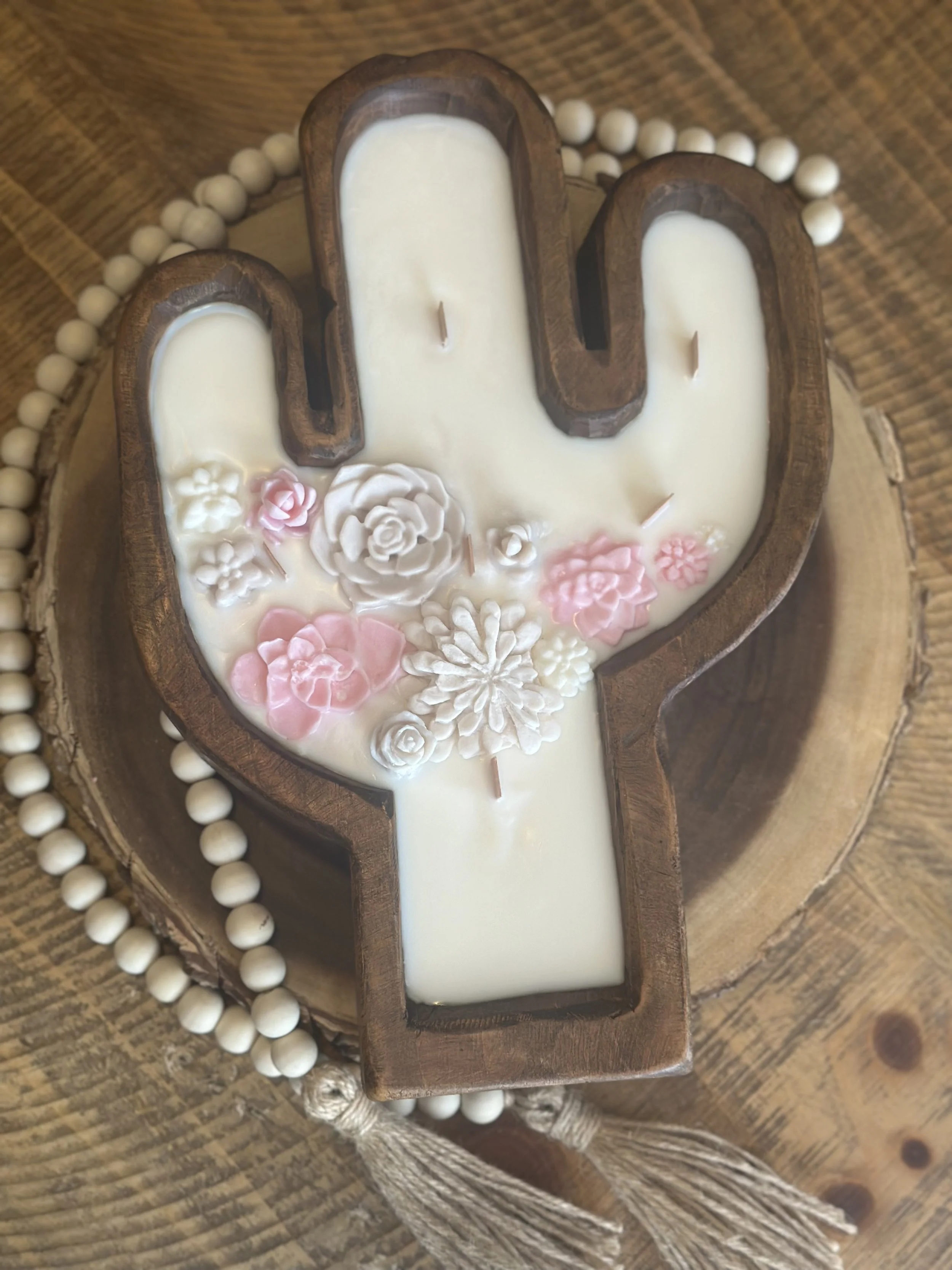 A candle shaped like a hand making a peace sign, decorated with pink and white flowers in a wooden holder on a wooden surface, surrounded by a string of white beads.