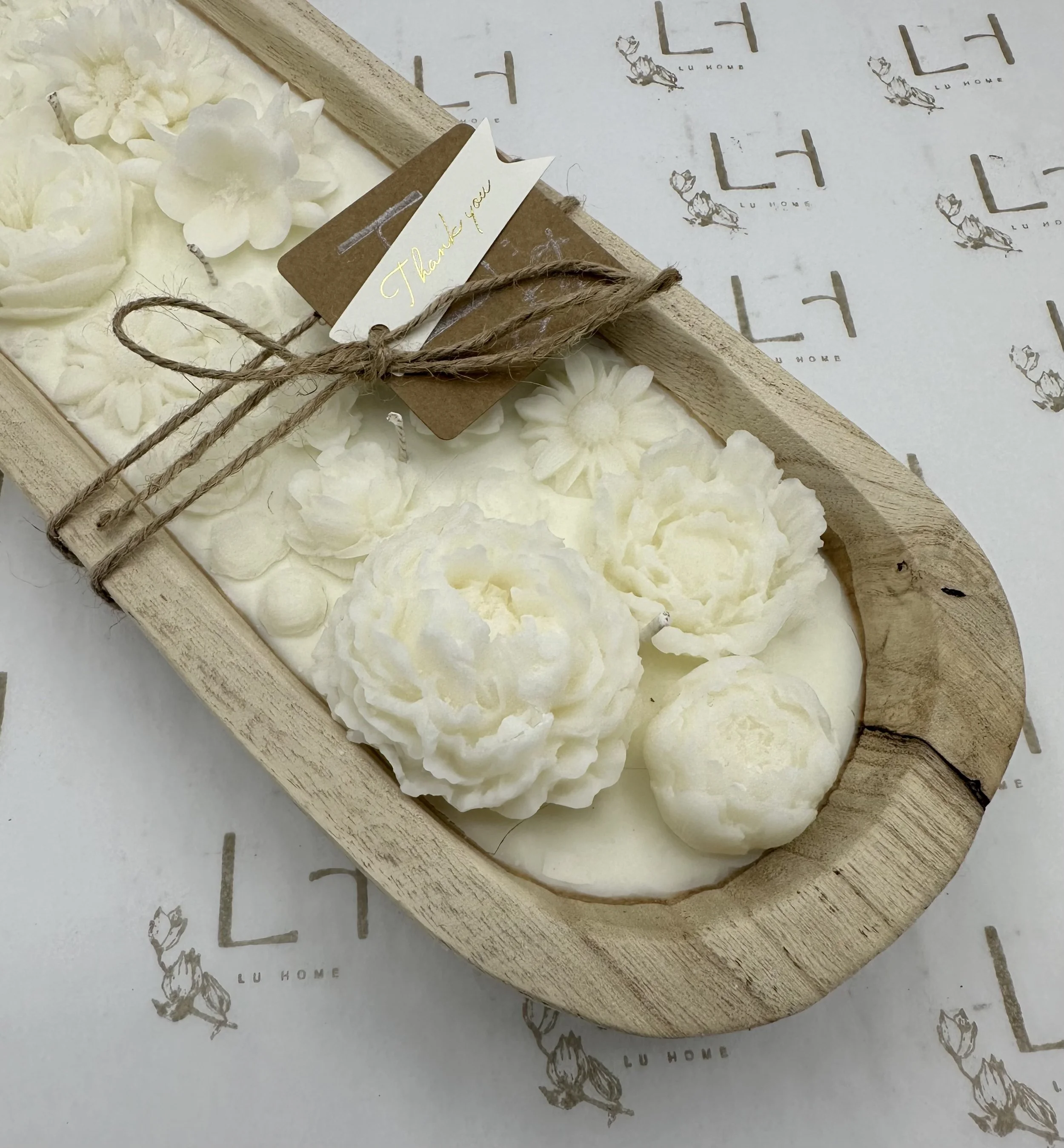 A wooden tray filled with white decorative soap flowers and spheres, tied with a string and a thank you tag, placed on paper with a logo pattern.