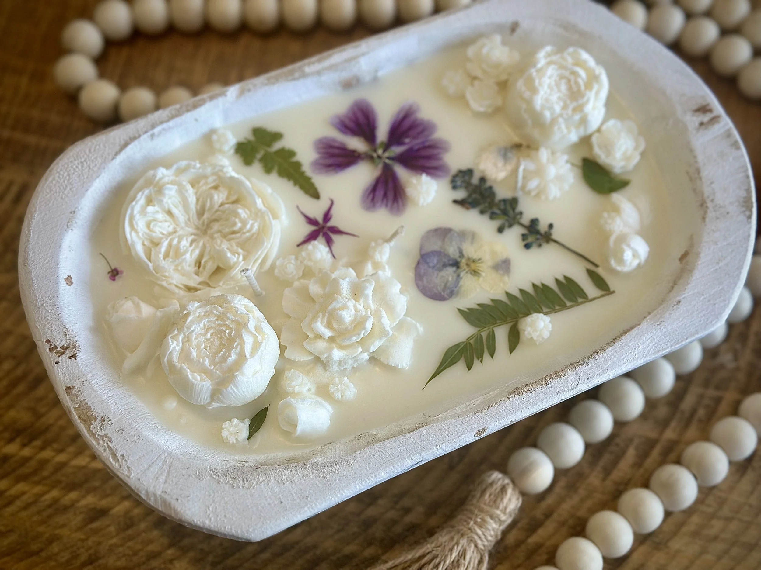 A soap bar with floral and botanical pressed flowers embedded on its surface, surrounded by a string of white beads, placed on a wooden surface.
