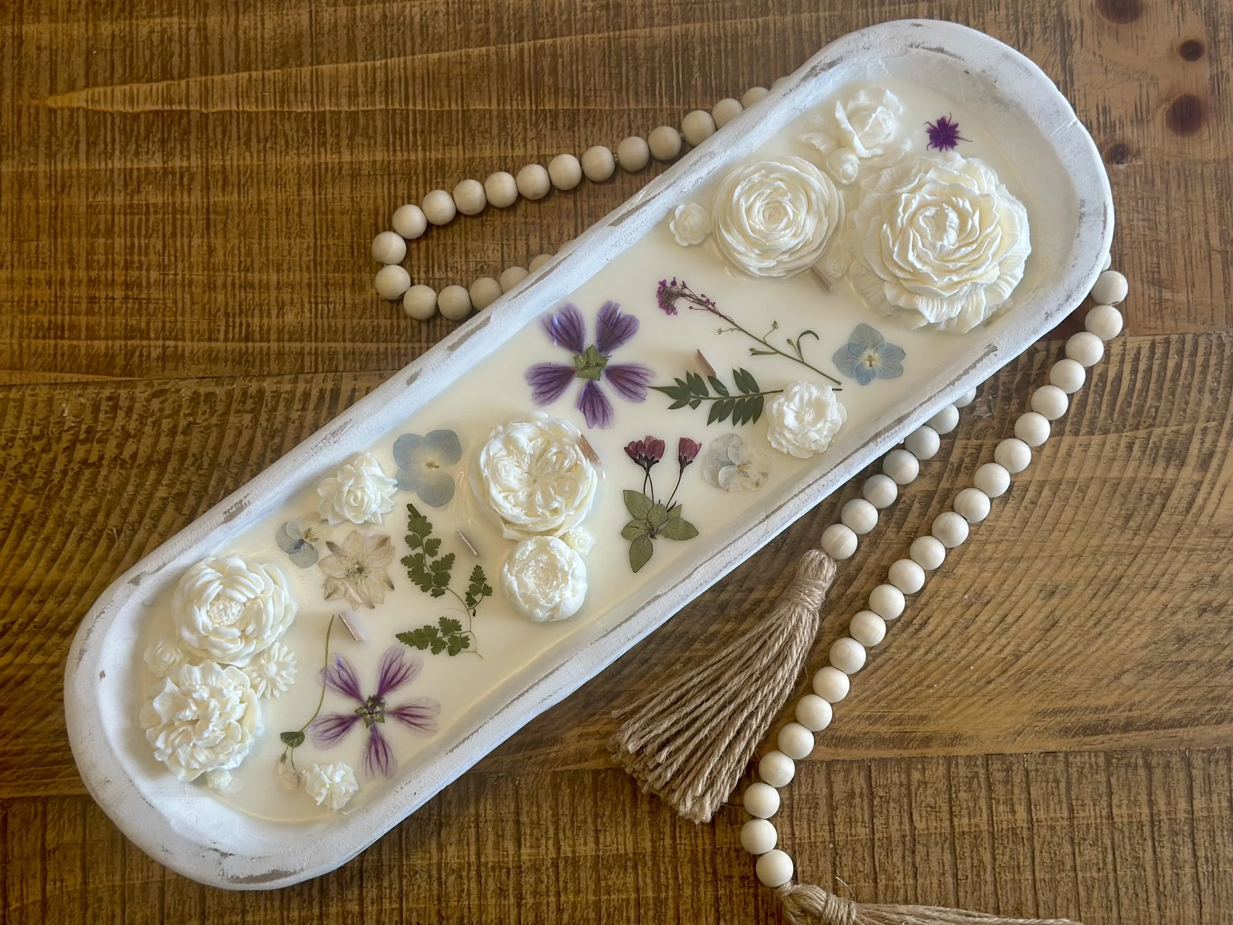 A white rectangular tray with decorative soap flowers and pressed real flowers, placed on a wooden surface, accompanied by a string of white beads and a brown tassel.