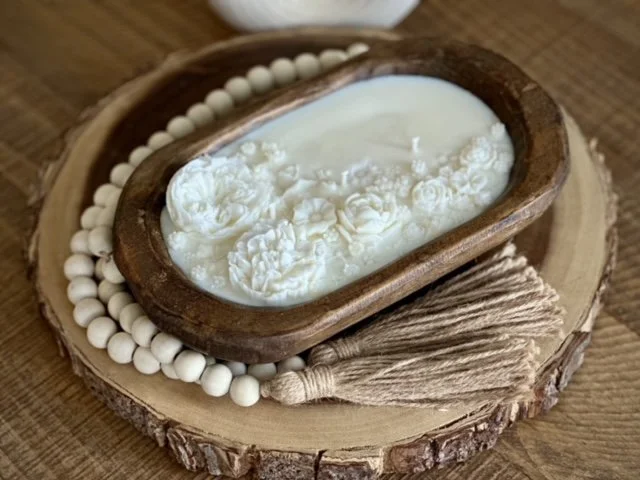 A wooden bowl with white decorative flowers inside, placed on a wooden slice with a bead necklace and a tassel decoration.