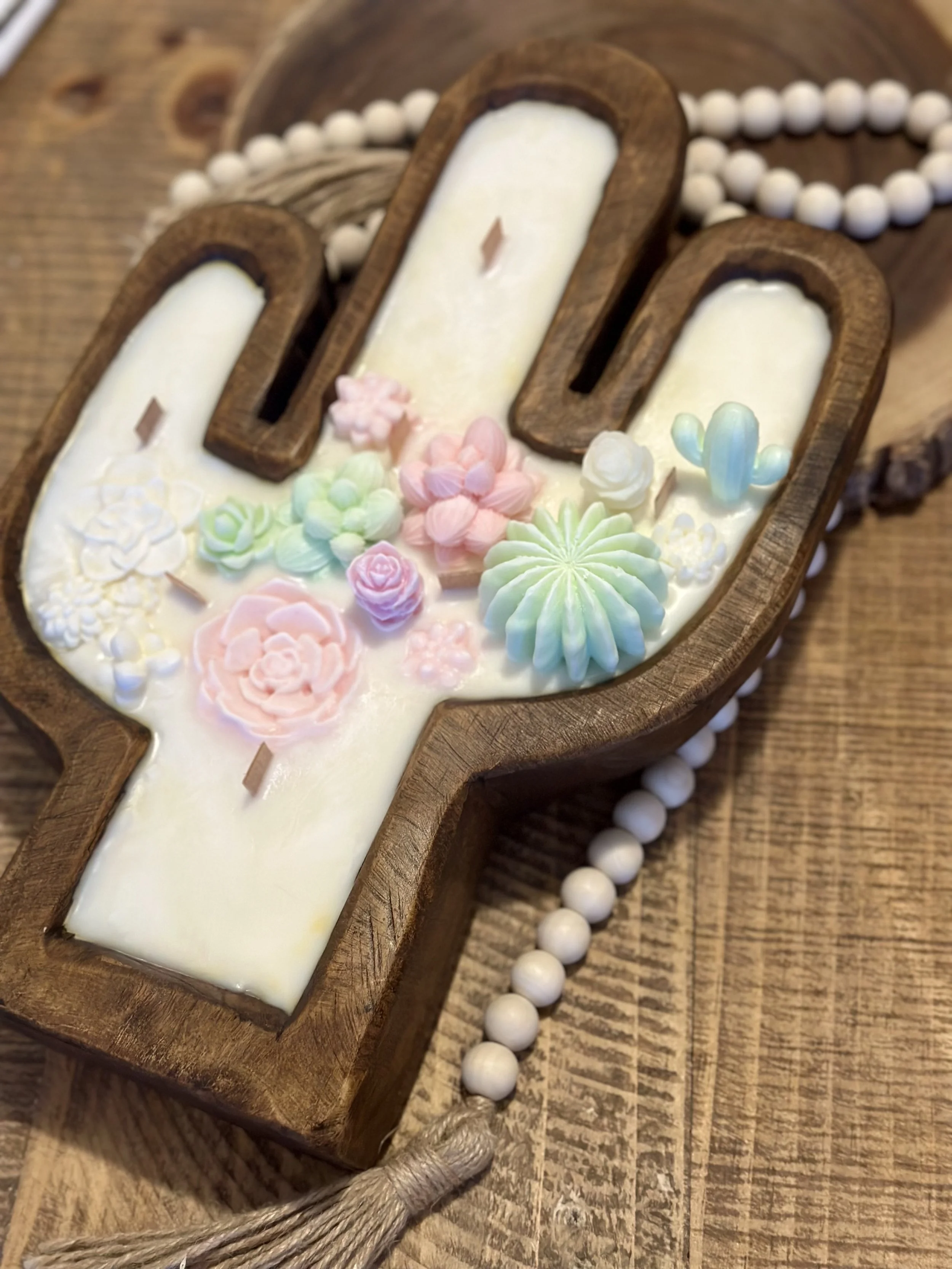 A candle shaped like a cross with a wooden border, decorated with pastel-colored flowers and a small cactus, placed on a wooden surface with a strand of white beads nearby.
