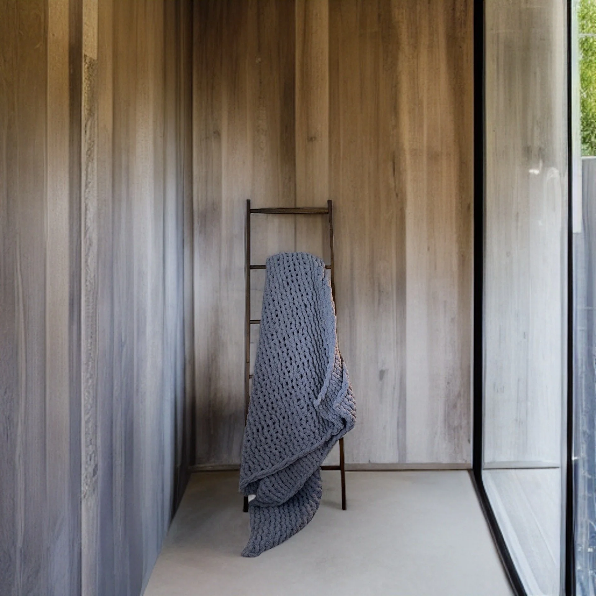 A minimal interior space with wood-paneled walls, a metal ladder with a gray knitted blanket hanging on it, and a large glass window showing greenery outside.