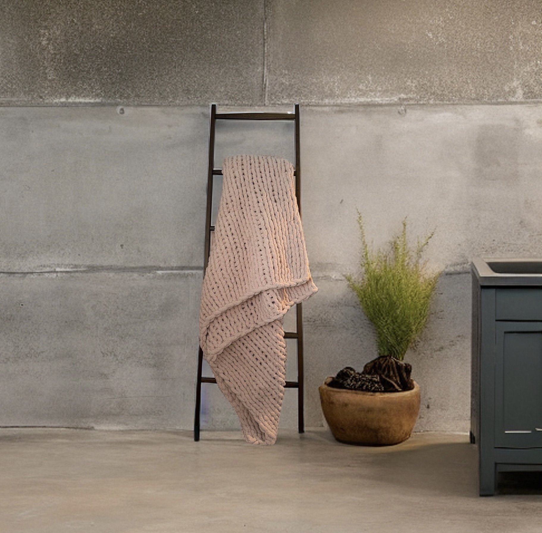 A pink knitted blanket draped over a black stepladder, with a potted plant with green feathery foliage next to it against a gray concrete wall.