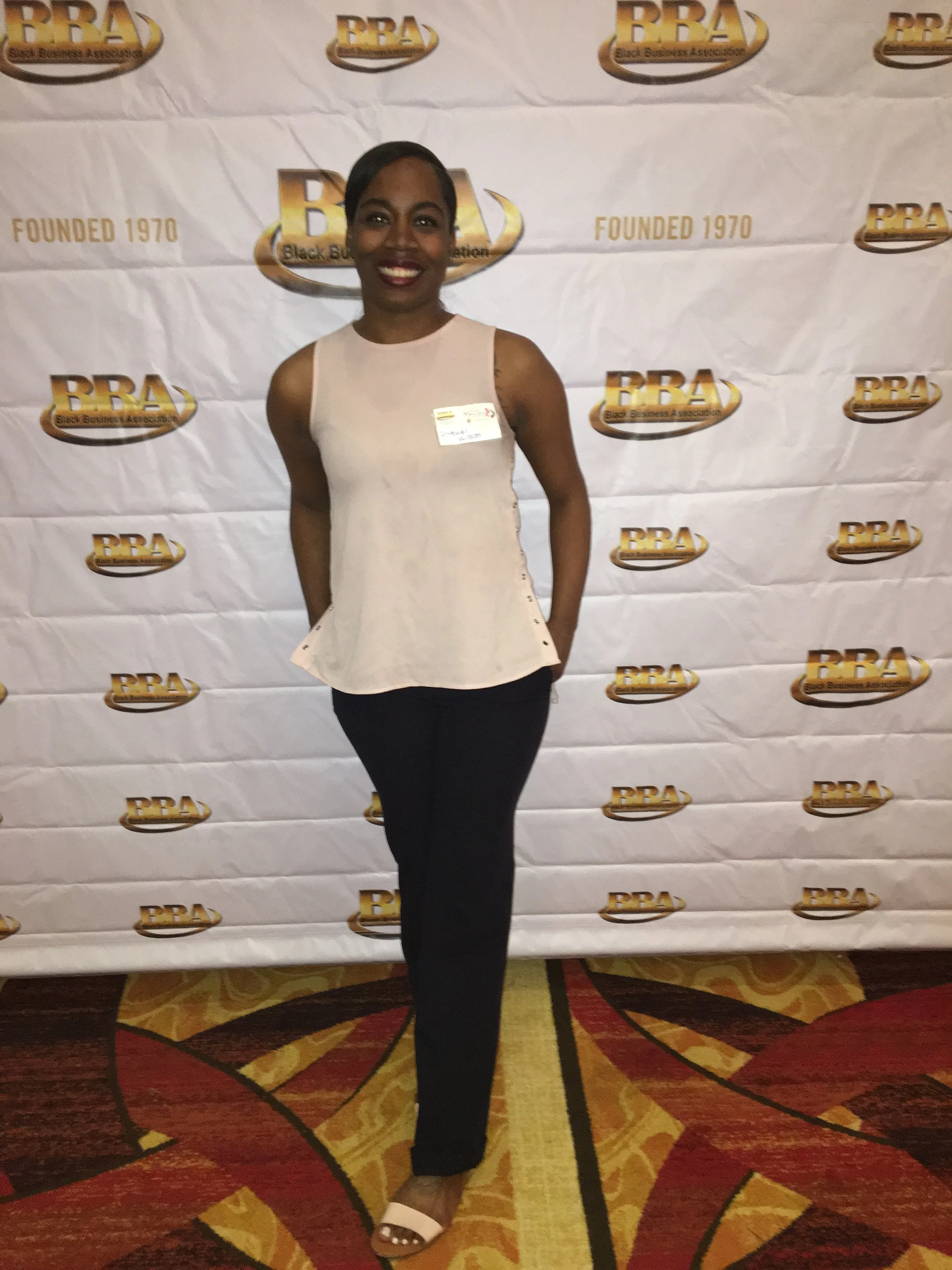 A woman standing in front of a step and repeat banner with the logo 'BBA,' which stands for Black Business Association, and the words, 'Founded 1970.' She is smiling, wearing a sleeveless beige top, black pants, and white open-toe shoes, with her right arm behind her back.