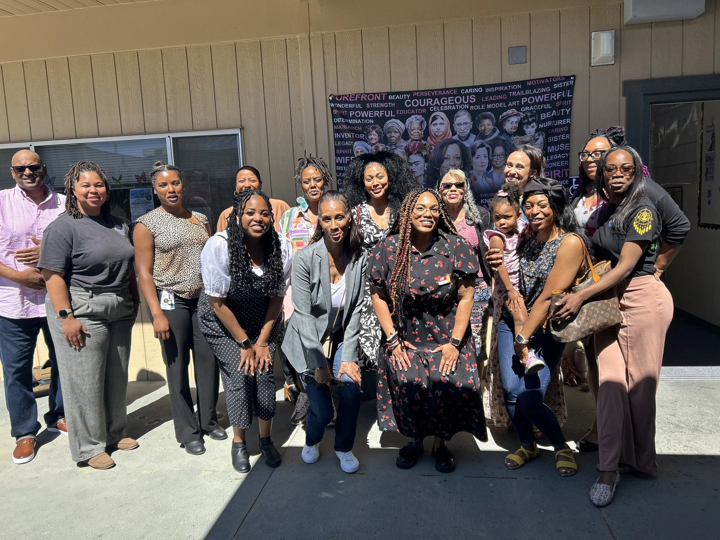 A diverse group of women and one man standing outdoors in front of a mural celebrating women. They are smiling and dressed in casual to business casual attire.