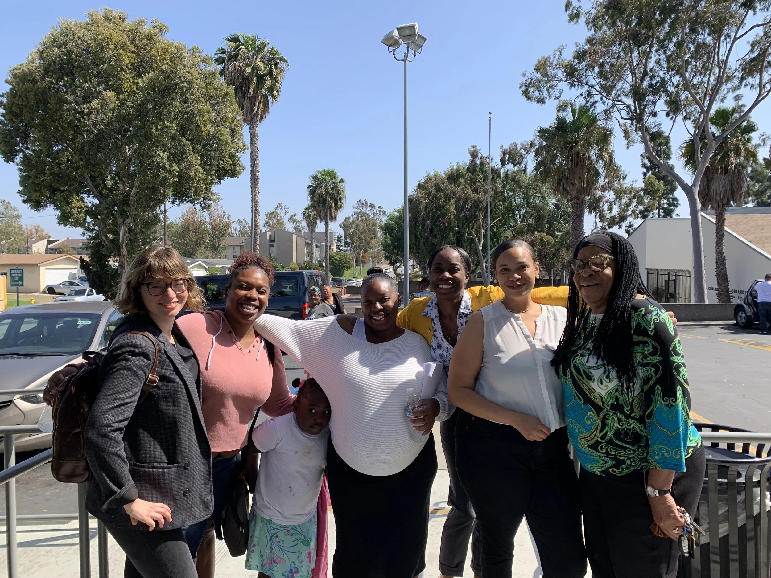 A group of six women and one young girl standing outdoors on a bright sunny day. They are smiling and posing for the camera with trees, parked cars, and buildings in the background.