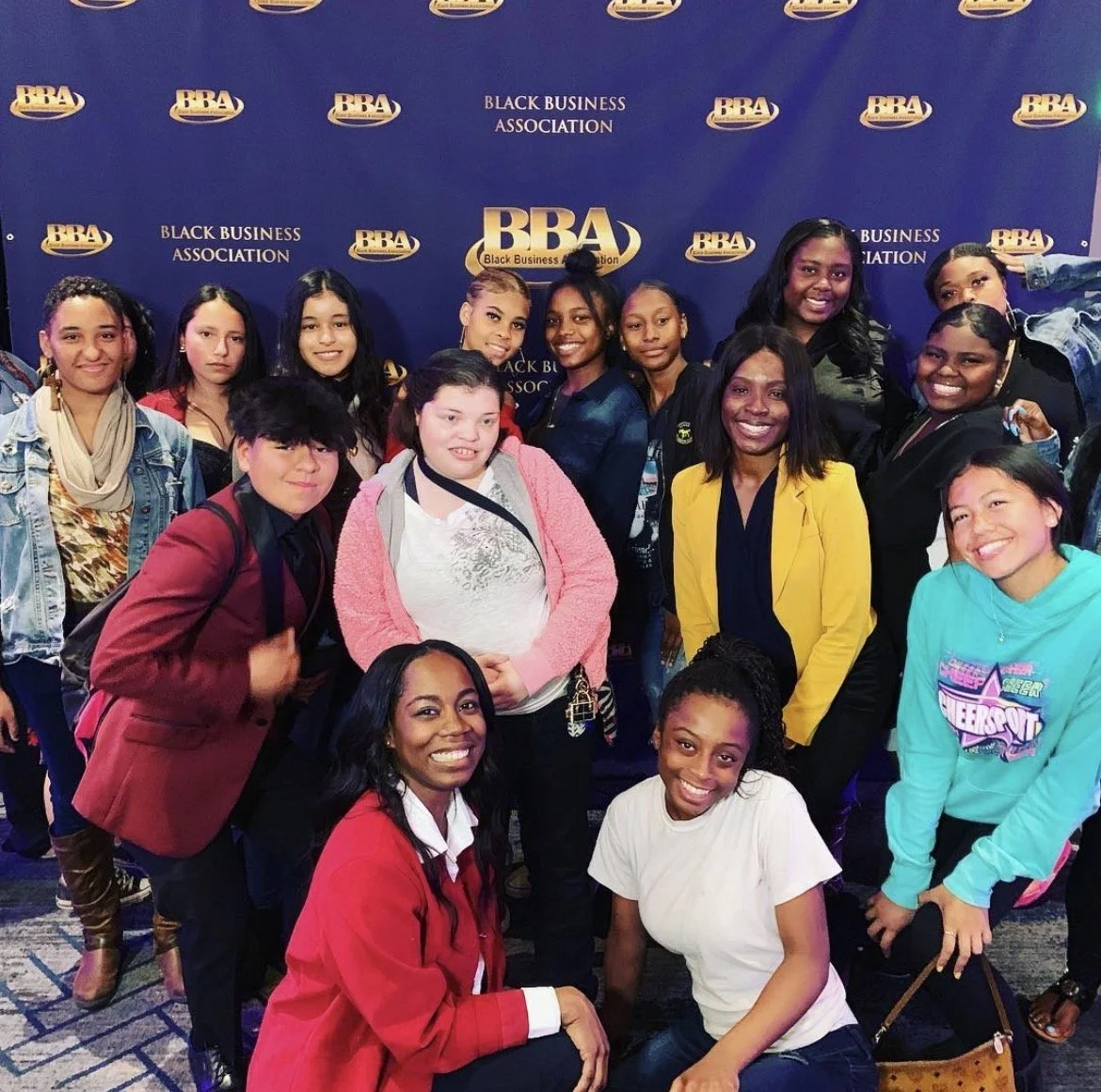 Group of diverse women and girls posing together at a Black Business Association event, standing in front of a blue backdrop with the BBA logo.
